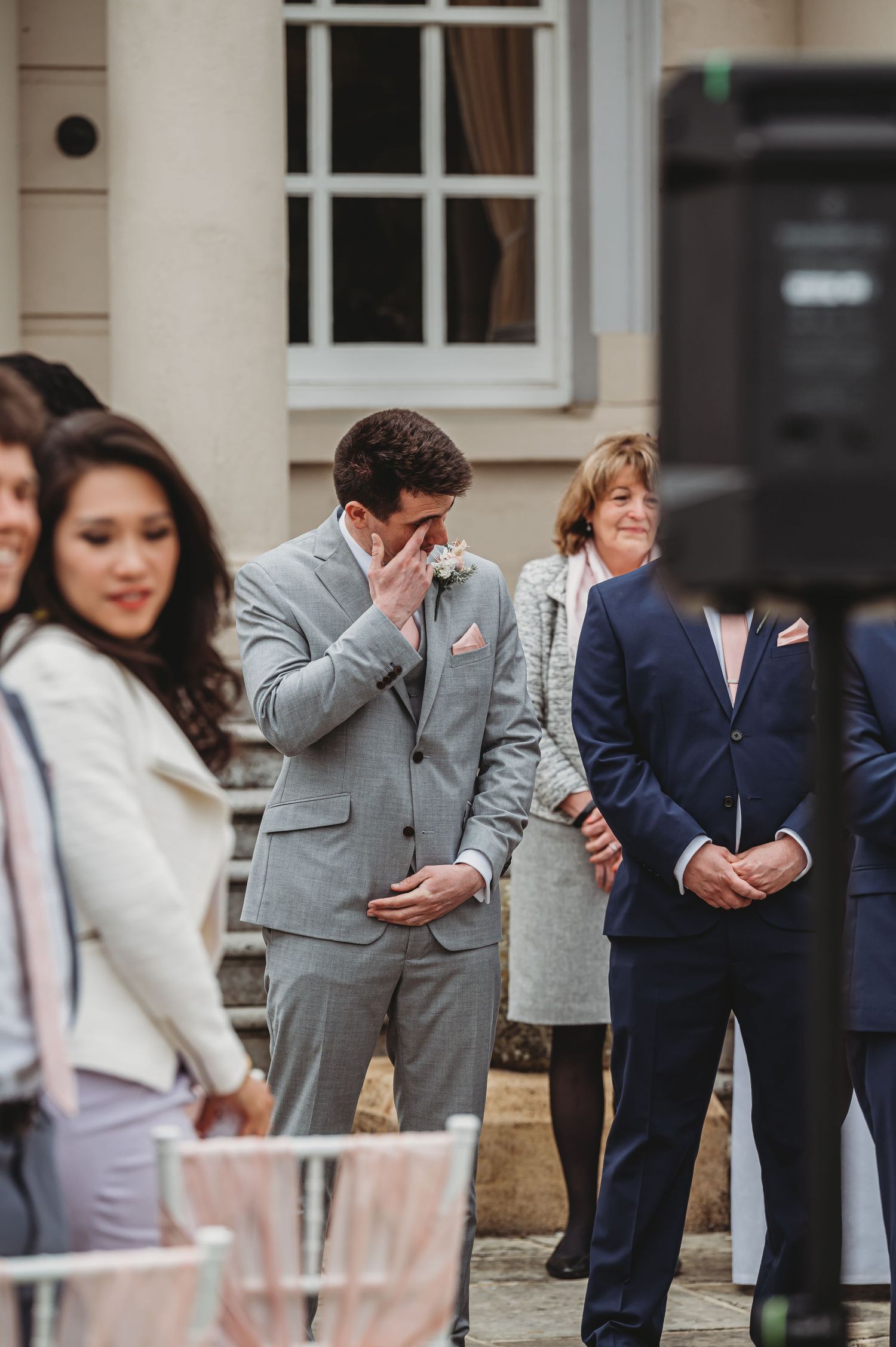An emotional moment captured during a wedding ceremony with attendees standing together outdoors near a windowed entrance.