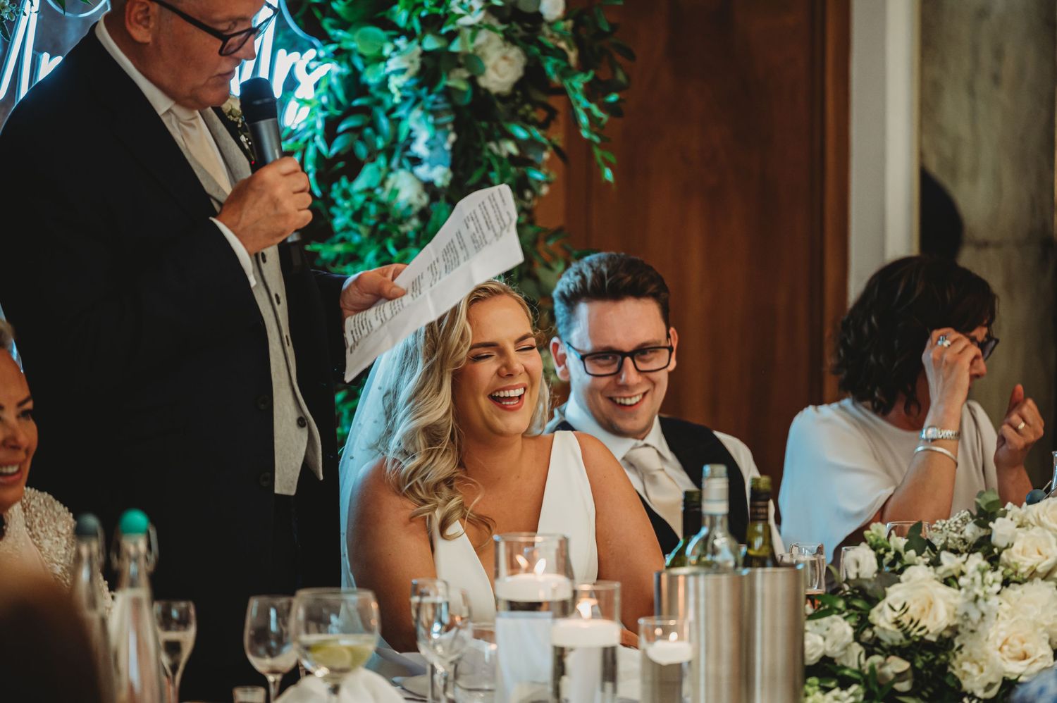 Wedding guests share laughter during reception dinner at an elegantly decorated table with wine glasses and floral centerpieces.