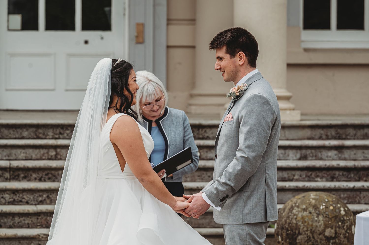 Wedding ceremony on stone steps with a bride in white gown, groom in gray suit, and officiant conducting the service.