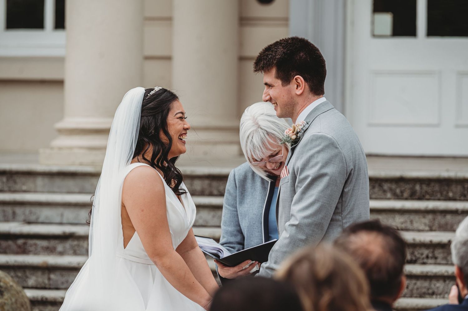 A couple shares a joyful moment during their wedding ceremony on stone steps outside a building with white columns.