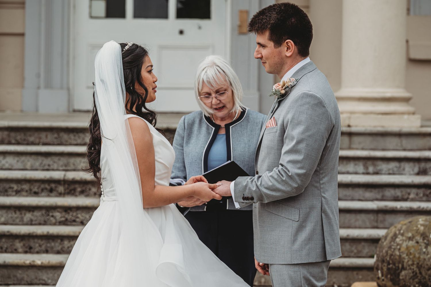 A couple exchanges wedding vows on stone steps while an officiant in black conducts their outdoor ceremony.