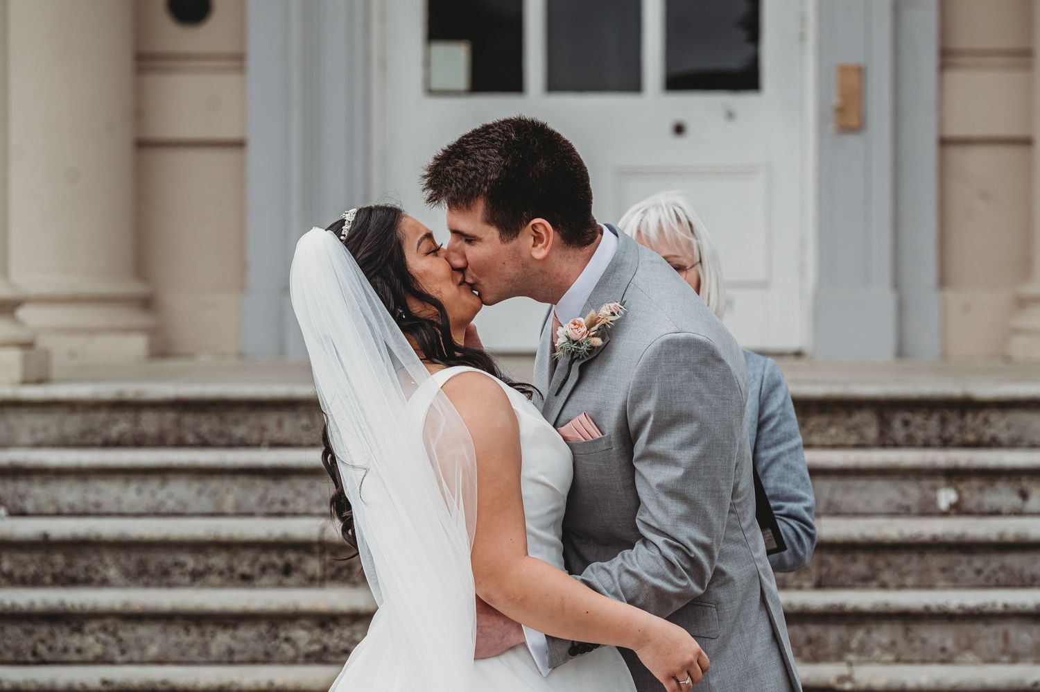 A bride in a white dress and groom in a gray suit share a romantic kiss on the steps of an elegant building.