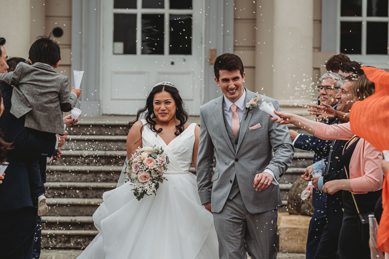 Newlyweds excitedly exit their wedding venue on stone steps while guests throw confetti in celebration.
