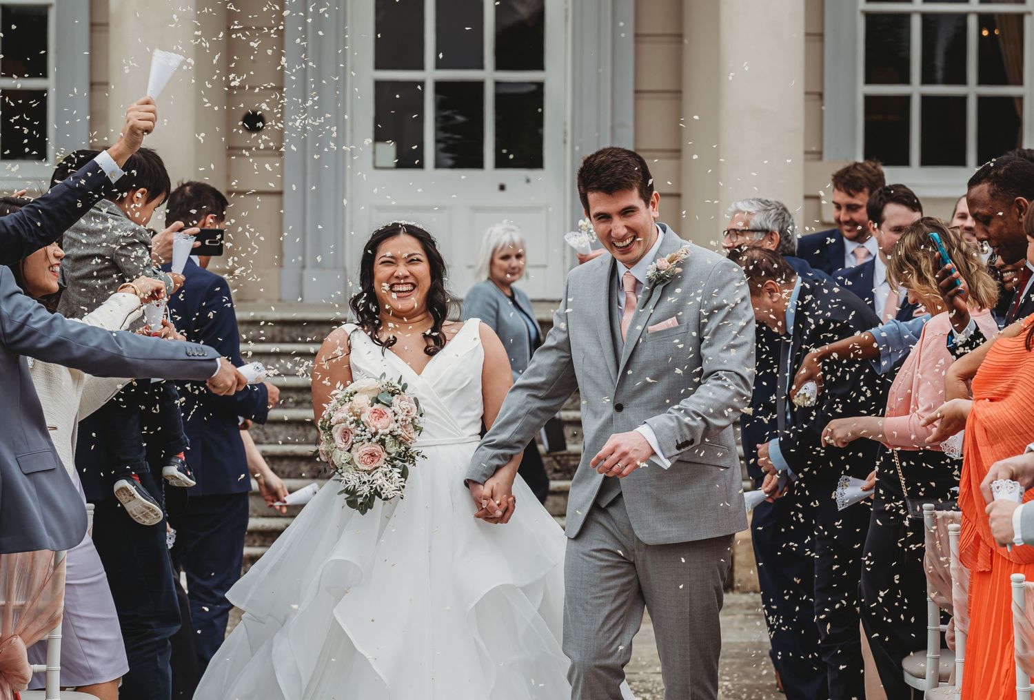 Happy newlyweds exit their wedding ceremony while guests shower them with confetti outside a historic building.