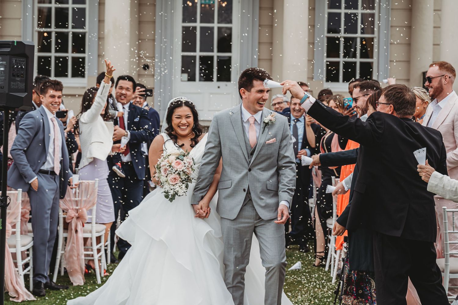 Wedding guests throw confetti over a newlywed couple exiting a historic building with white columns and windows.