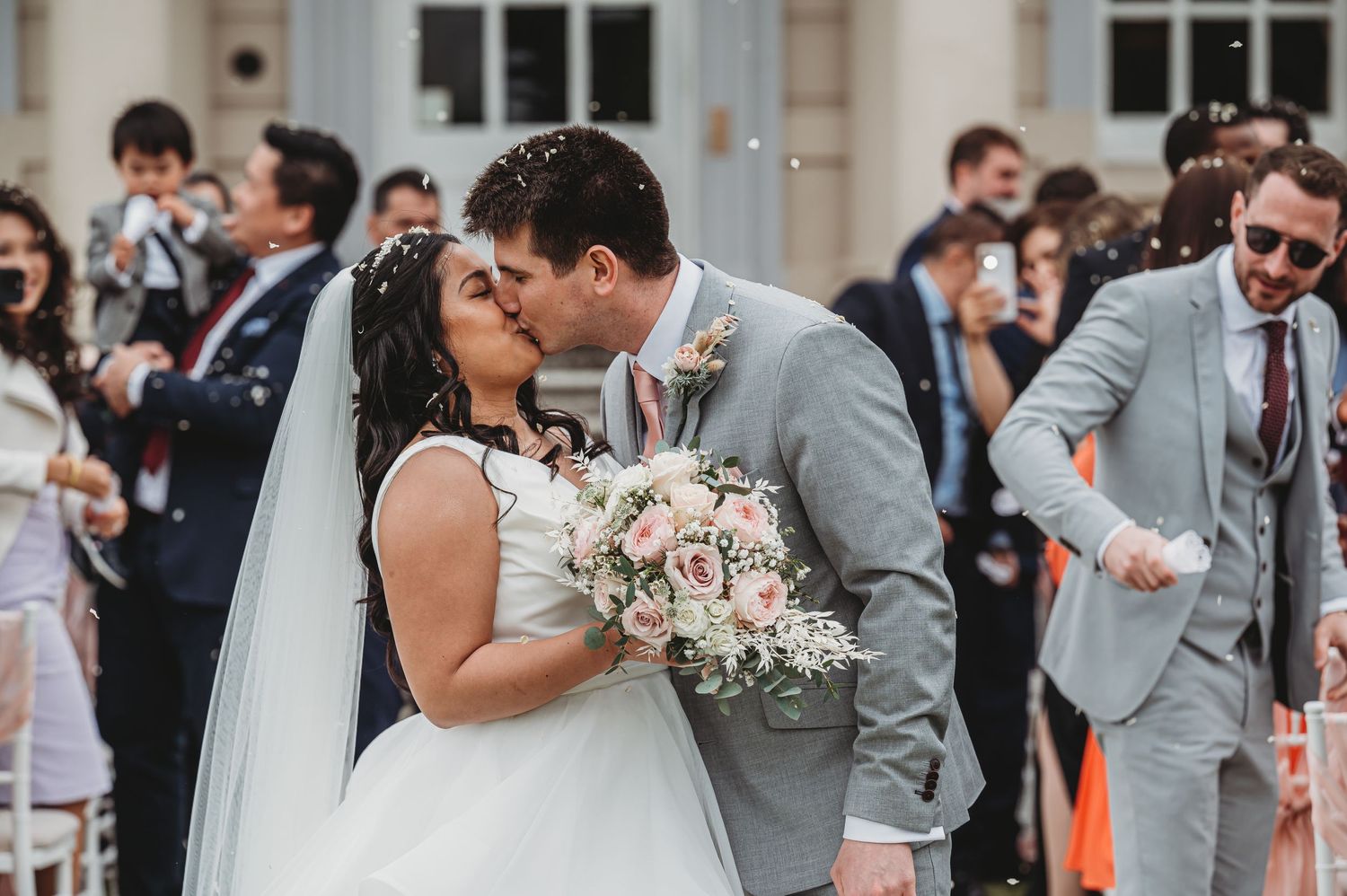 A joyous wedding couple shares a kiss while holding a blush pink and white bouquet surrounded by guests.