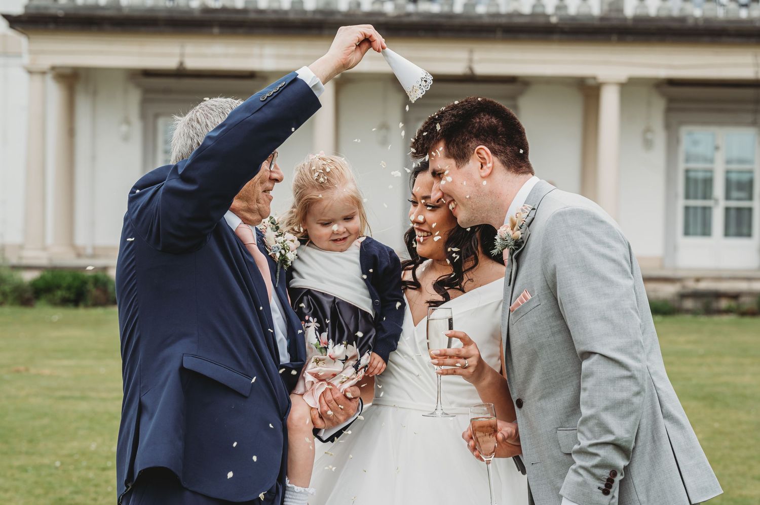 Wedding party celebrates with confetti on a green lawn in front of classic building.