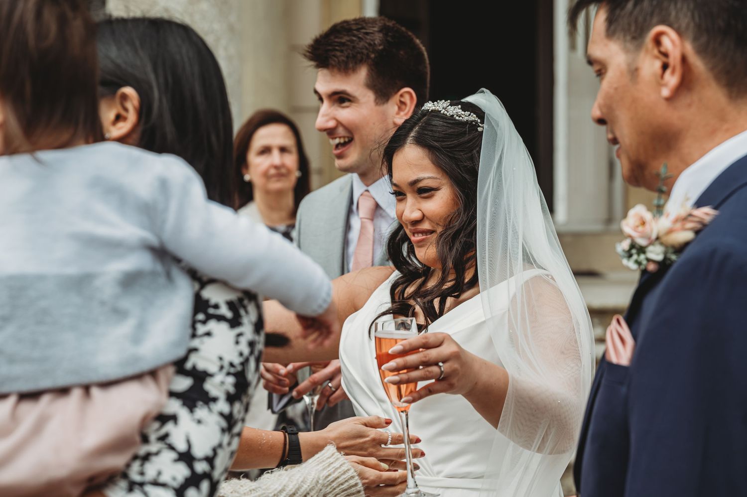 Happy newlyweds in formal wedding attire receive congratulations from guests outside during their celebration.