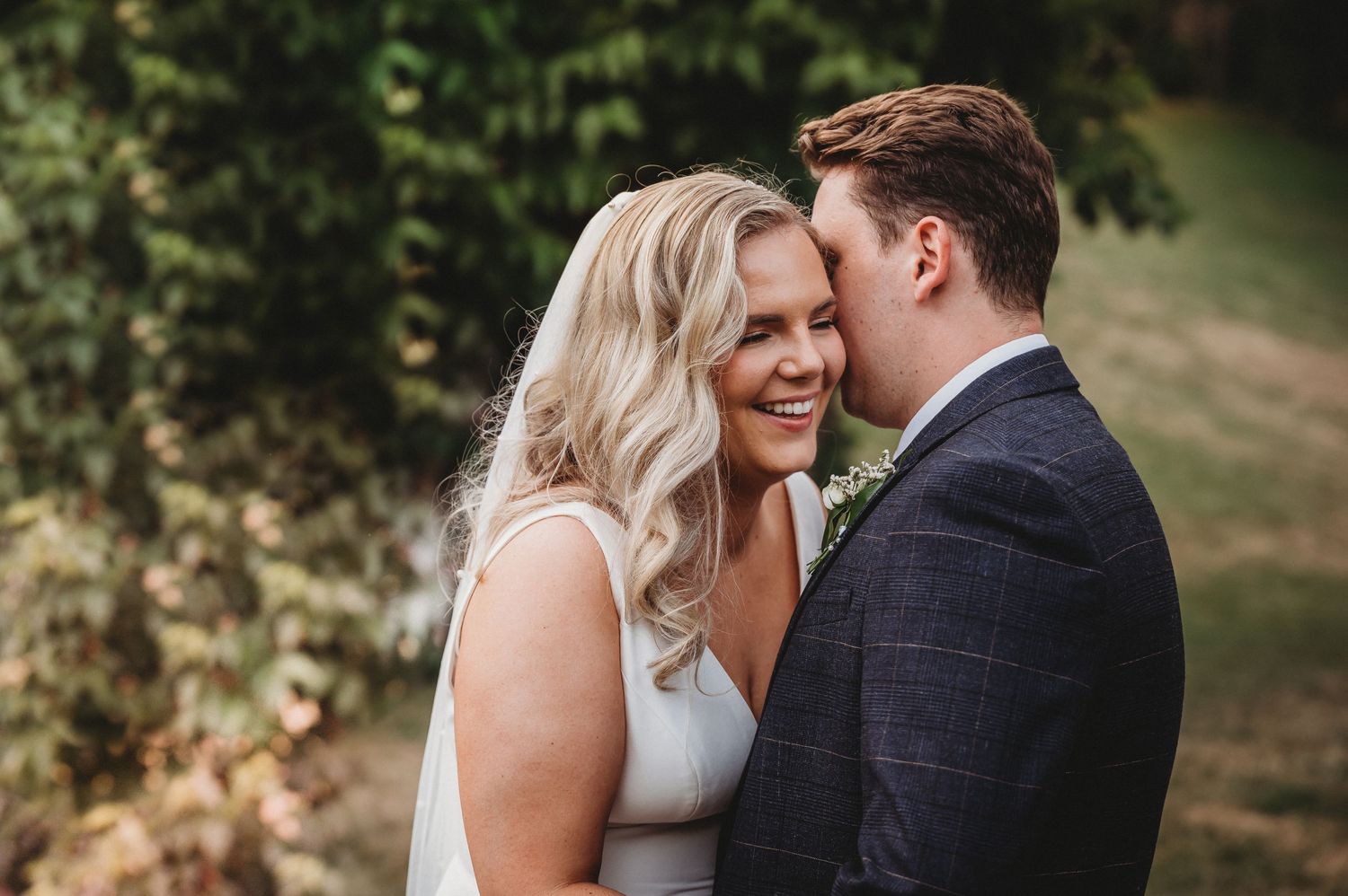 A romantic couple shares an intimate moment outdoors, with the bride wearing white and groom in navy plaid suit.