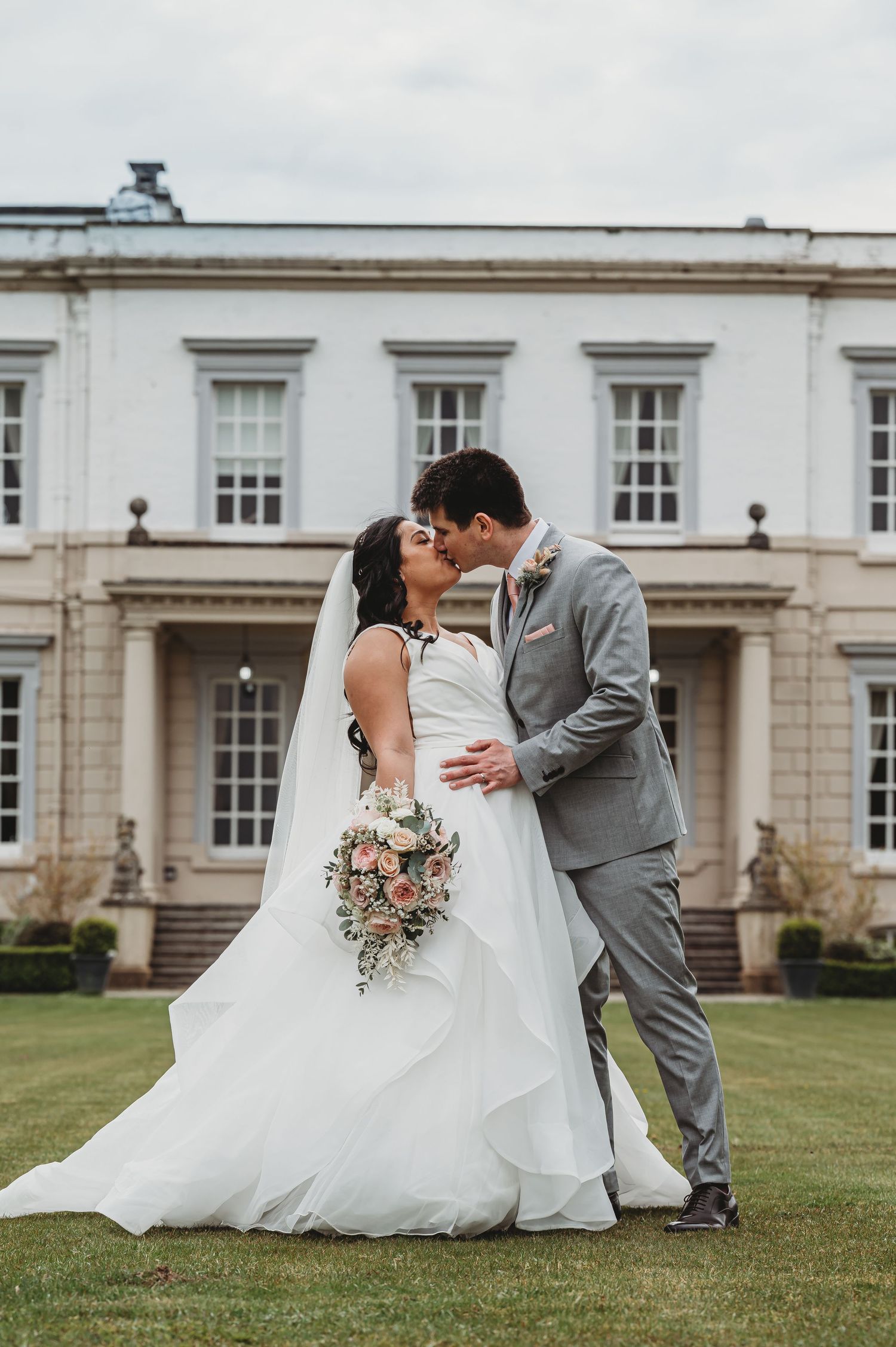 Newlyweds share a romantic kiss on the lawn of an elegant white mansion with a bouquet of pink flowers.