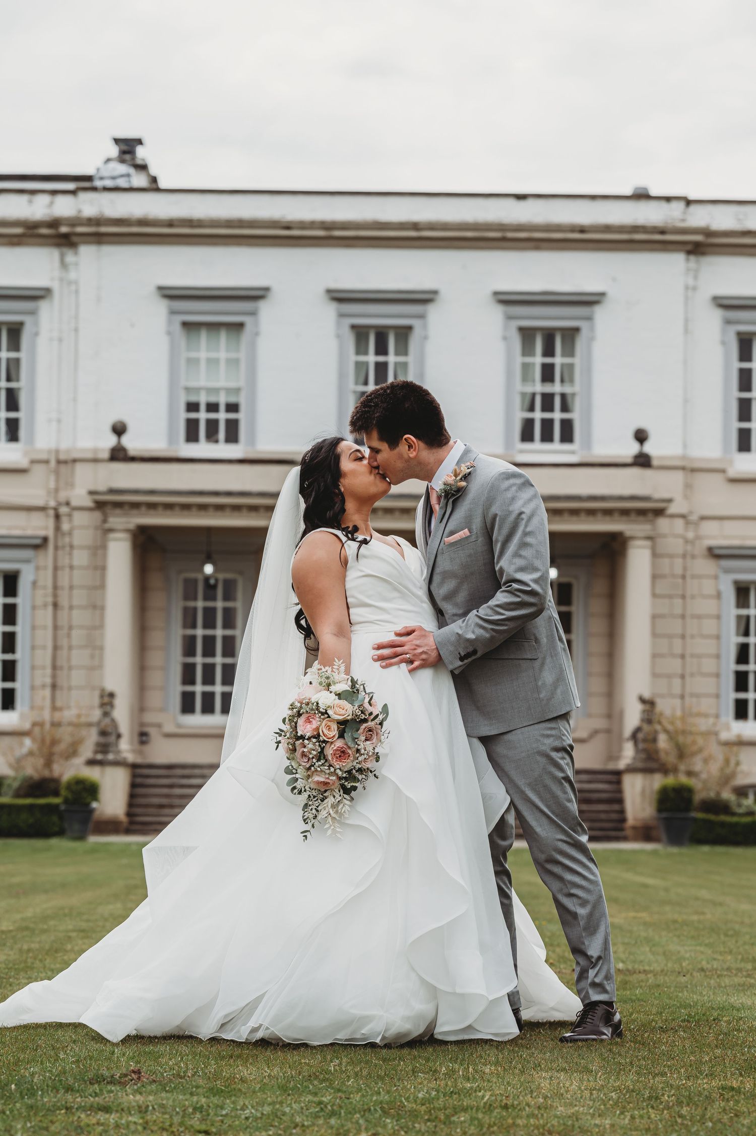 A romantic wedding couple shares a tender kiss on the lawn of an elegant mansion with white columns and Georgian architecture.