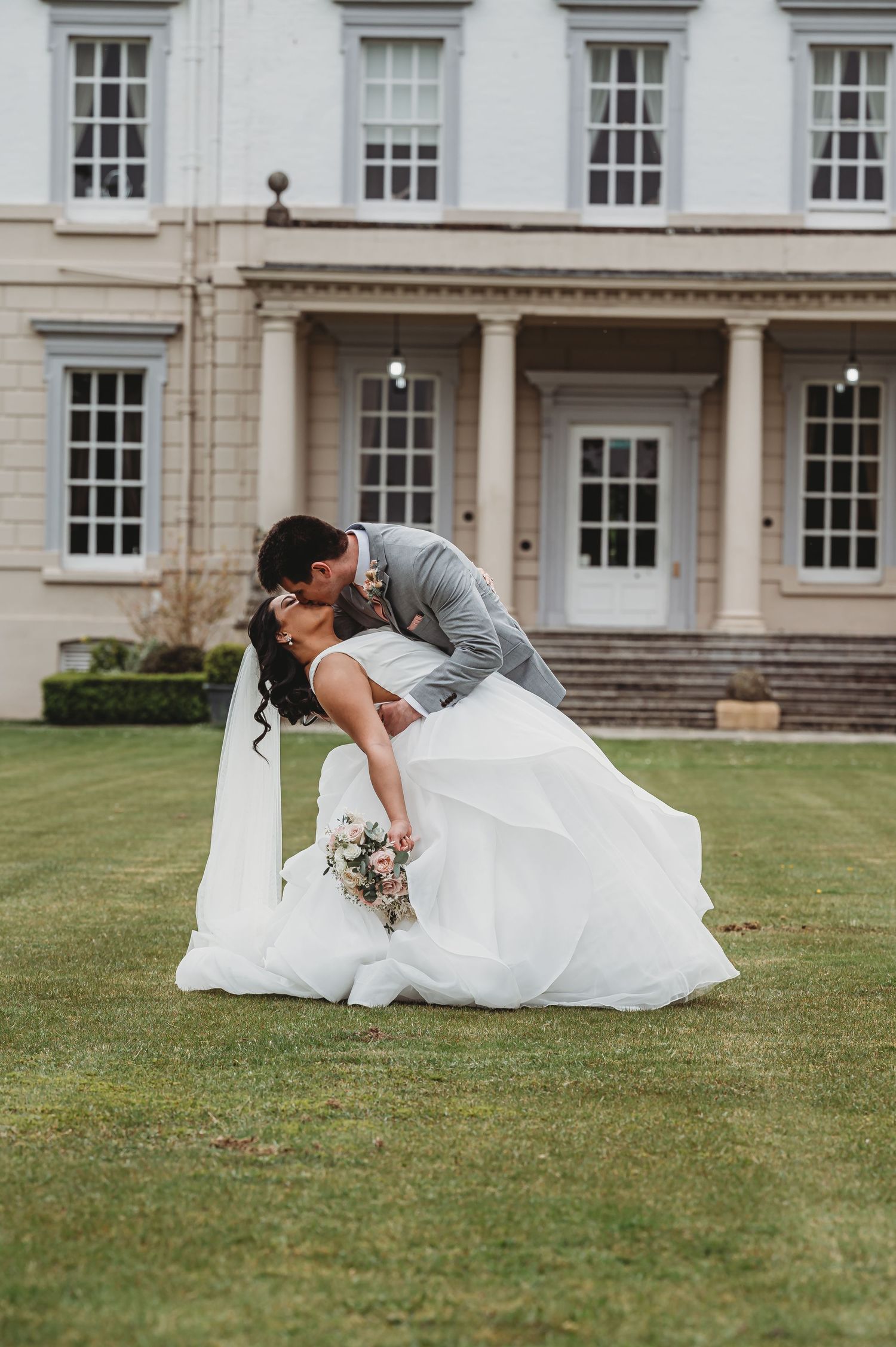 Newlyweds share romantic dip kiss on lawn in front of elegant white mansion during wedding photoshoot.