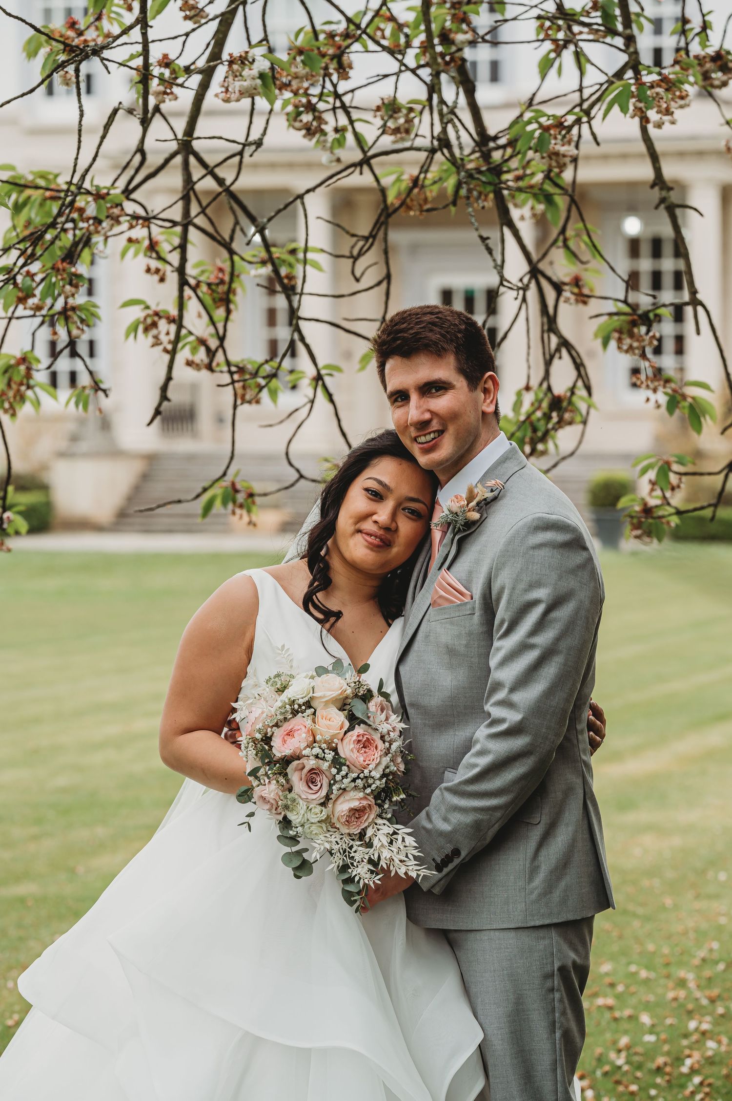 Wedding couple embraces under blooming tree branches while holding pink and white floral bouquet on manicured lawn.