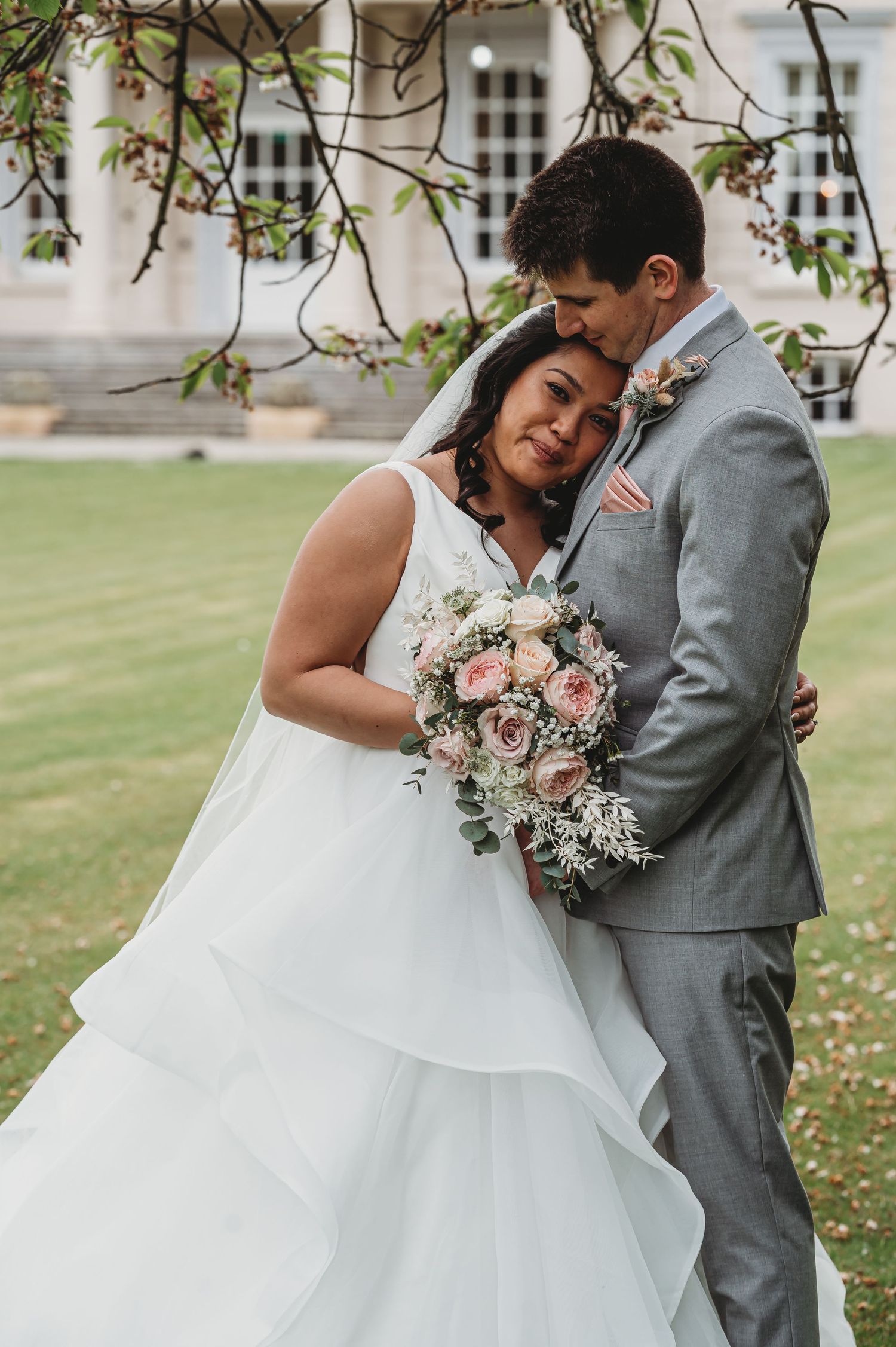 Newlyweds embrace outdoors with a stunning floral bouquet featuring pink and white flowers under blooming trees.