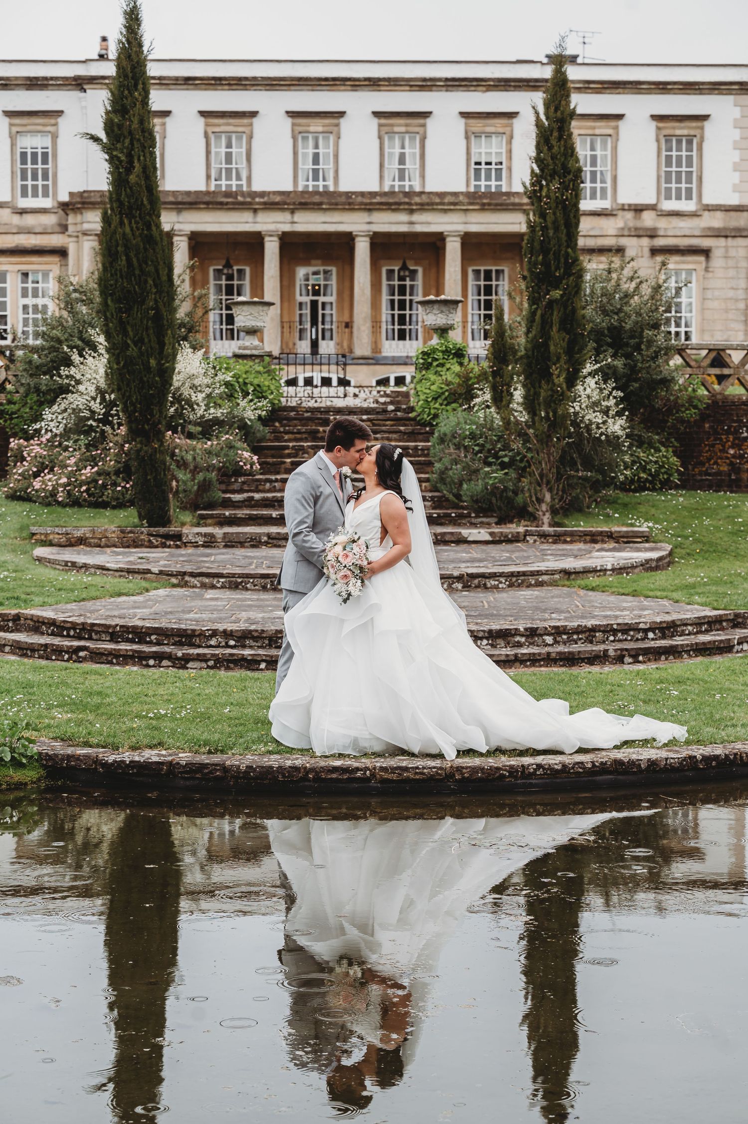A couple embraces at their wedding beside a reflective pond at an elegant manor house with tall cypress trees.