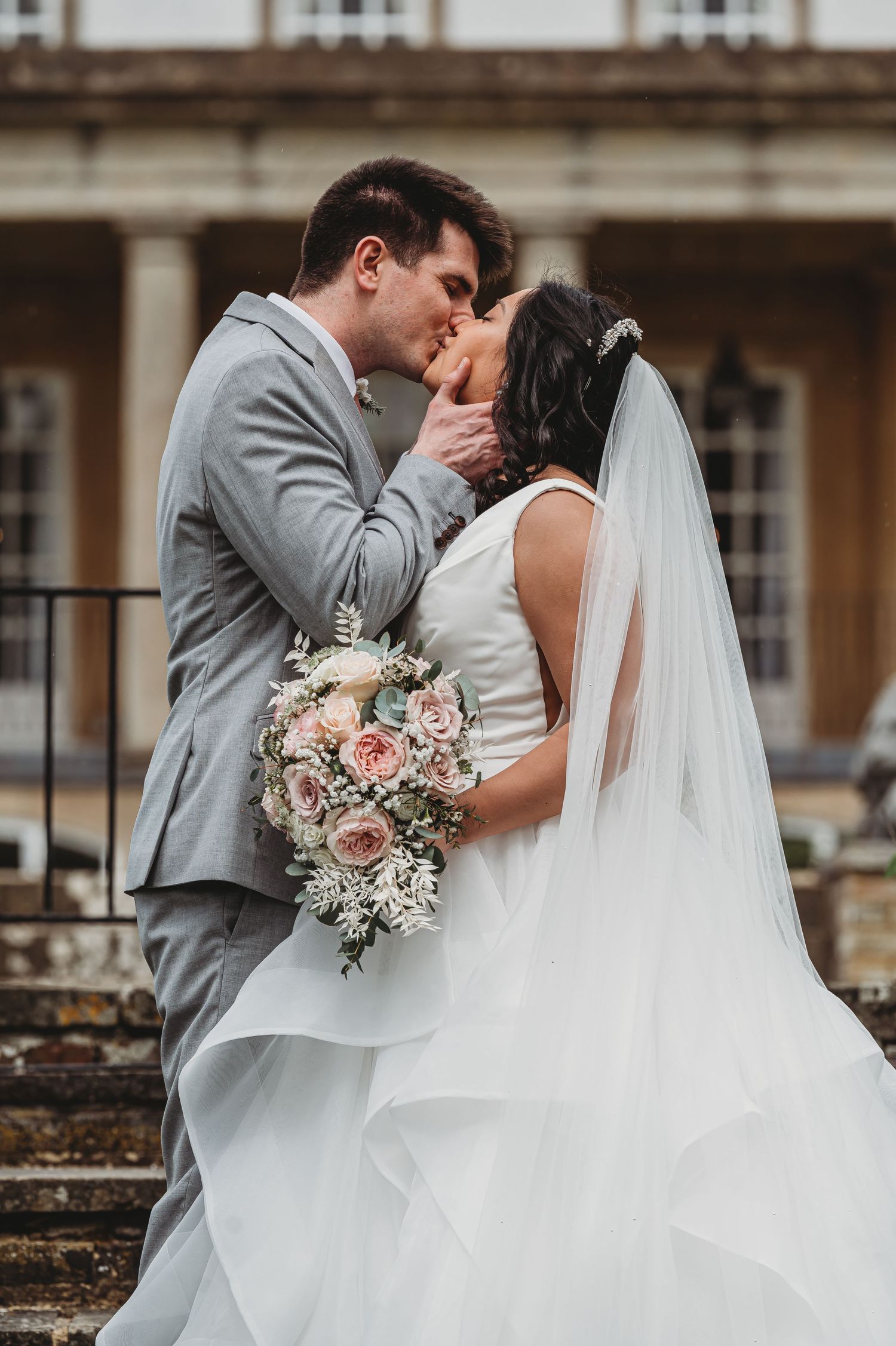 Bride holds blush pink and white bouquet during intimate wedding kiss on stone steps at historic venue.