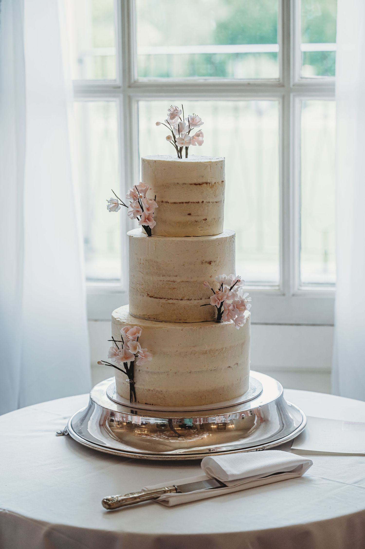 A four-tier semi-naked wedding cake with delicate pink floral accents sits elegantly by a bright window.
