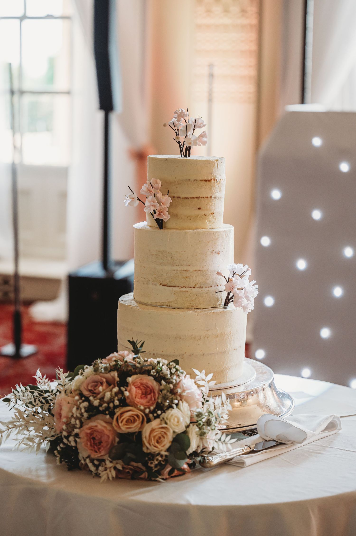 Elegant three-tiered white wedding cake decorated with delicate pink flowers and surrounded by a romantic rose bouquet.