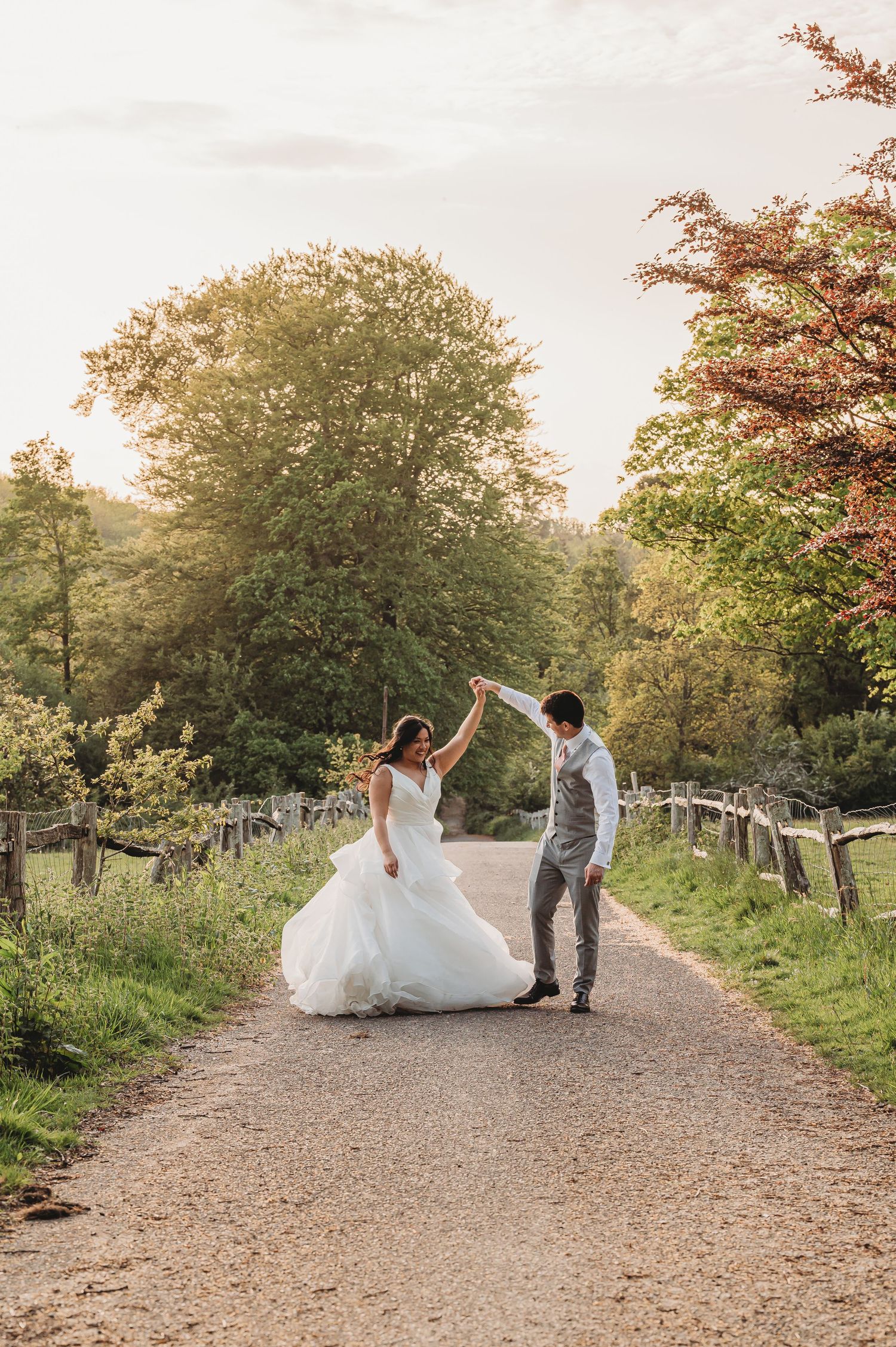 A couple dances on a tree-lined dirt path during their romantic countryside wedding photoshoot at sunset.