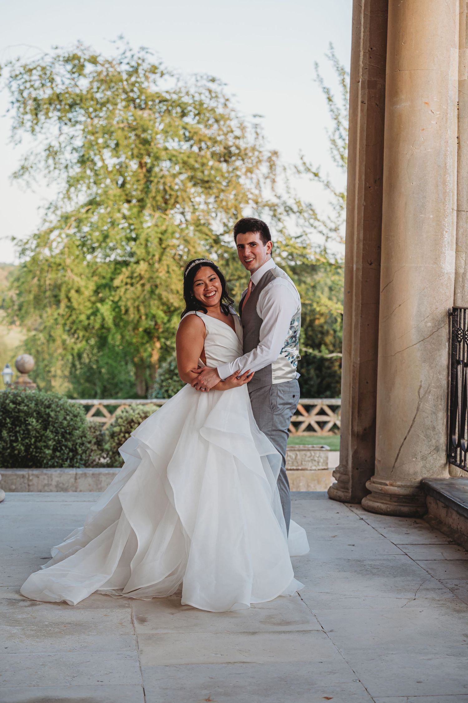 Newlywed couple embraces on stone patio with flowing white wedding dress and garden backdrop.