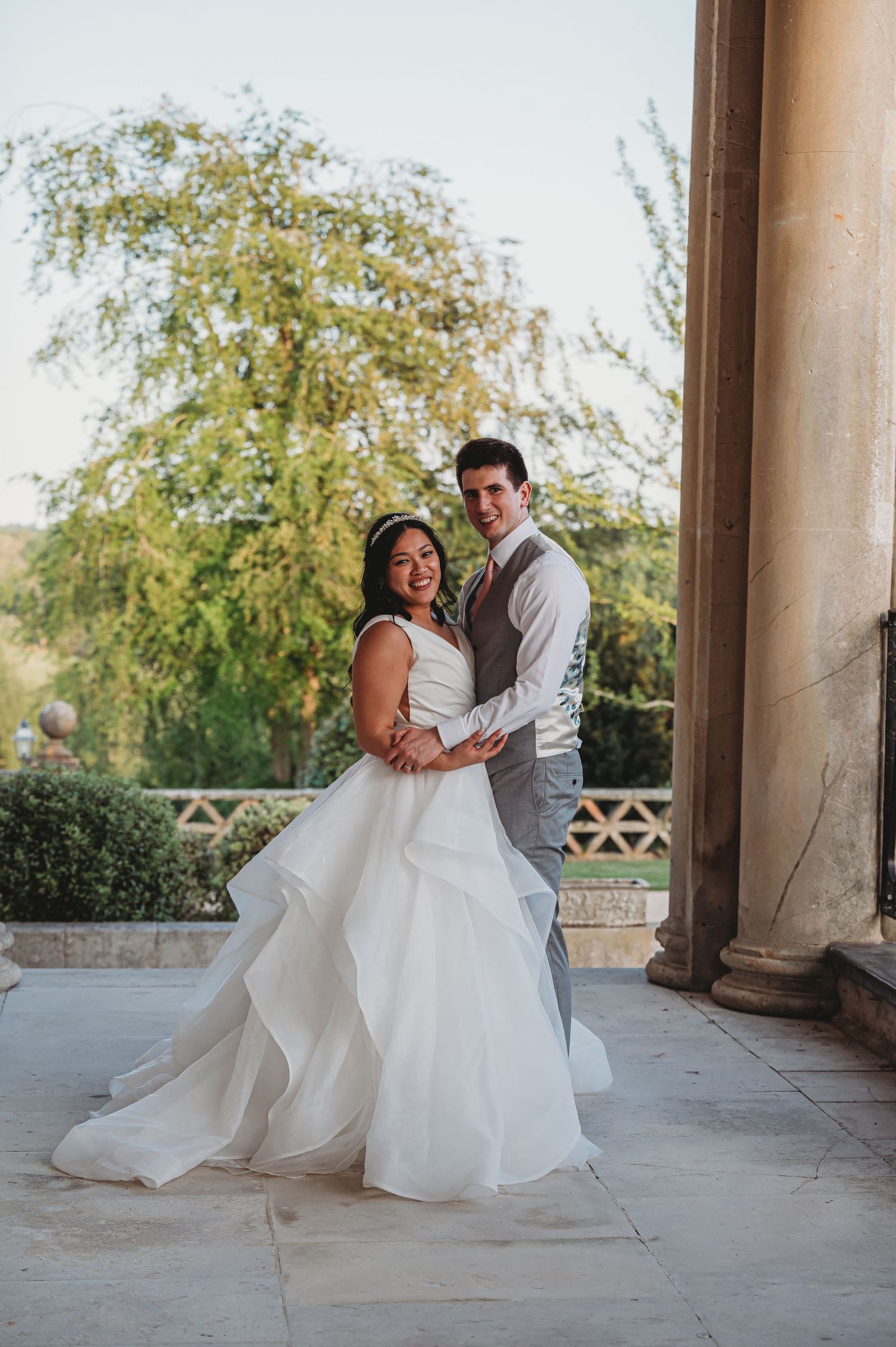 A couple poses together on a stone patio with flowering trees in the background during a romantic wedding photoshoot.