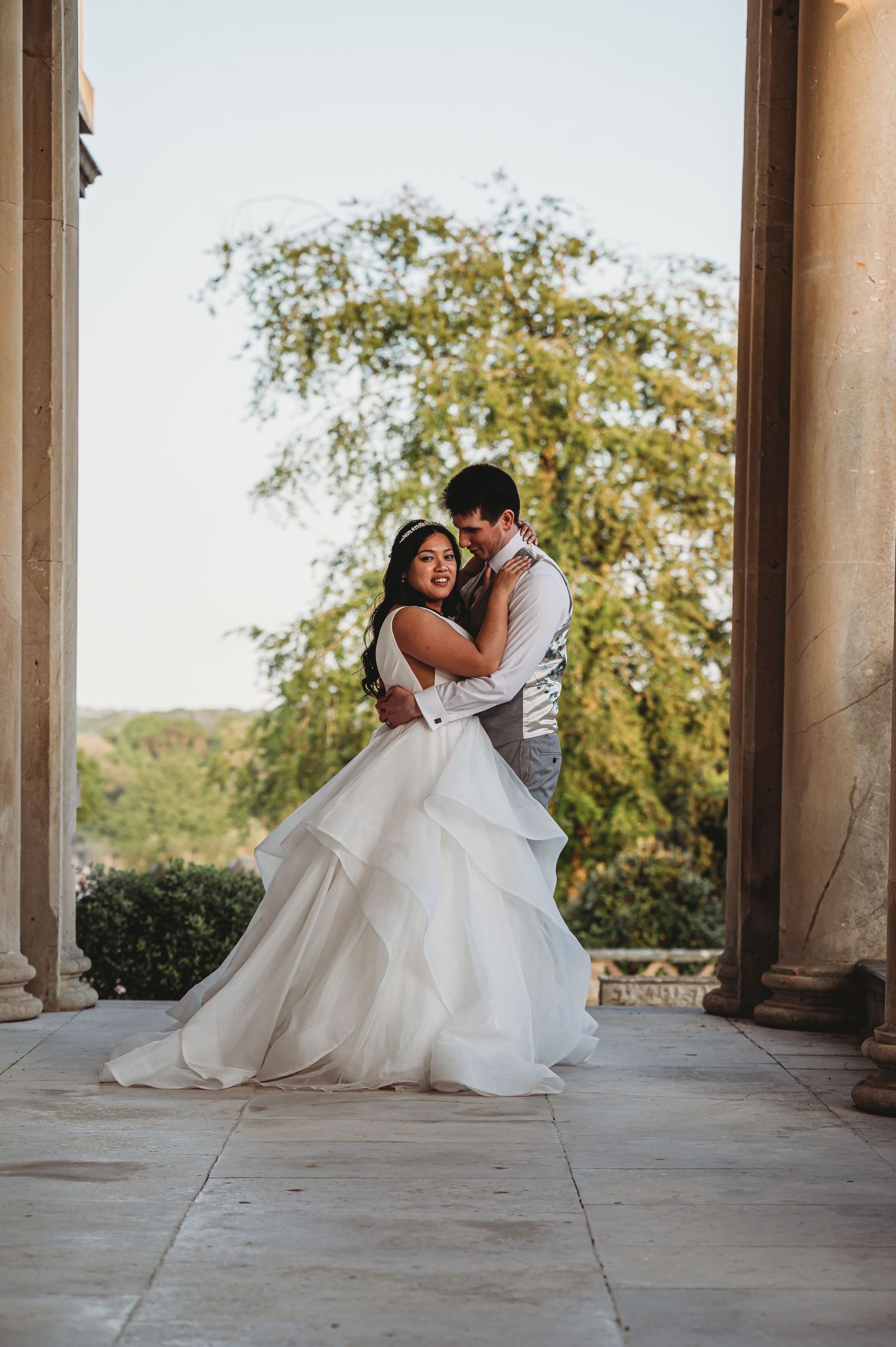 A romantic wedding couple embraces and dances together on a stone terrace with sunlit trees in the background.
