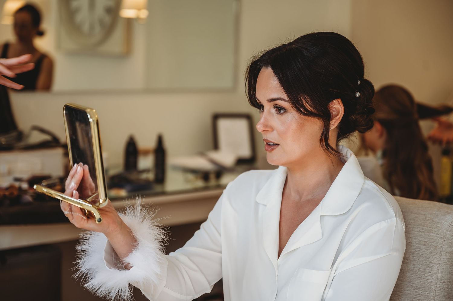 Person in white robe checking their makeup in a compact mirror during wedding preparations.