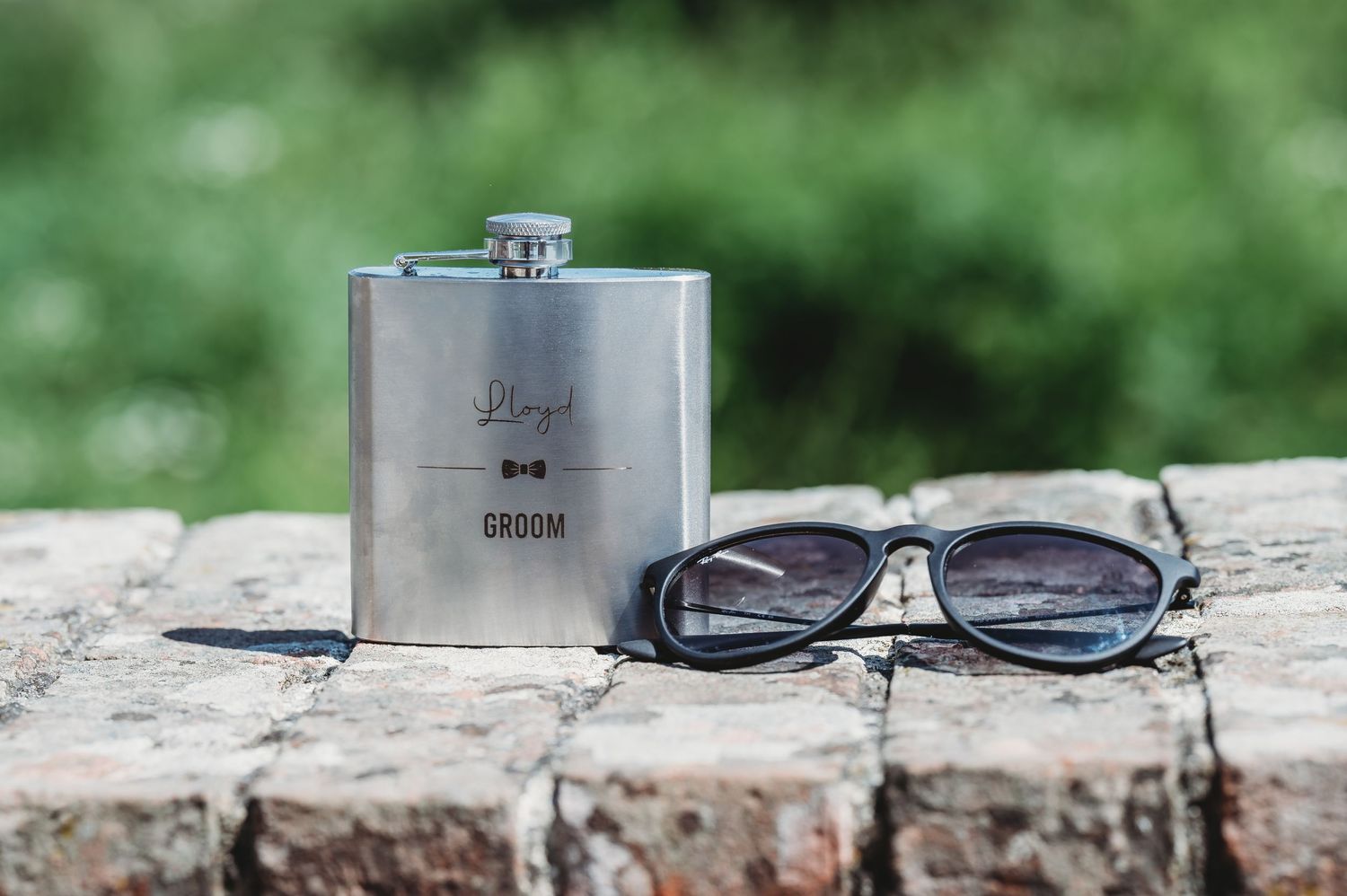 Metal hip flask and sunglasses arranged on rustic wooden surface outdoors.