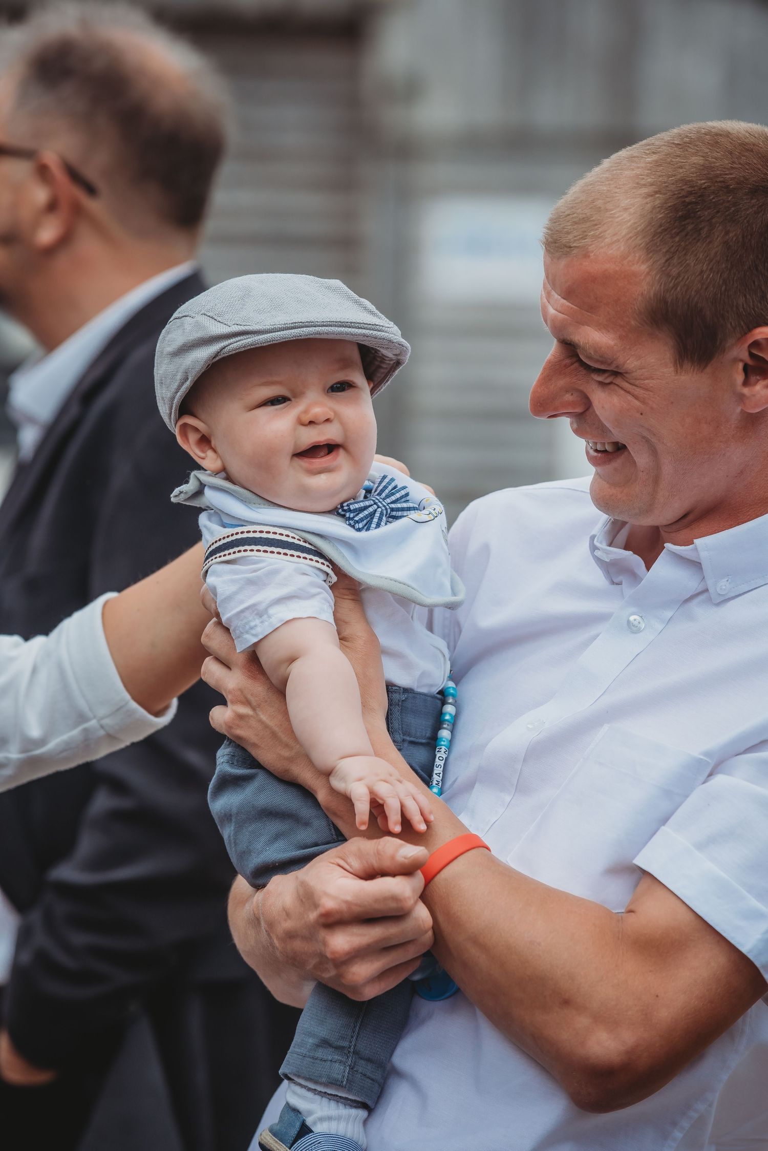 A smiling baby wearing a gray flat cap and blue outfit is lovingly held by an adult in a white button-down shirt.