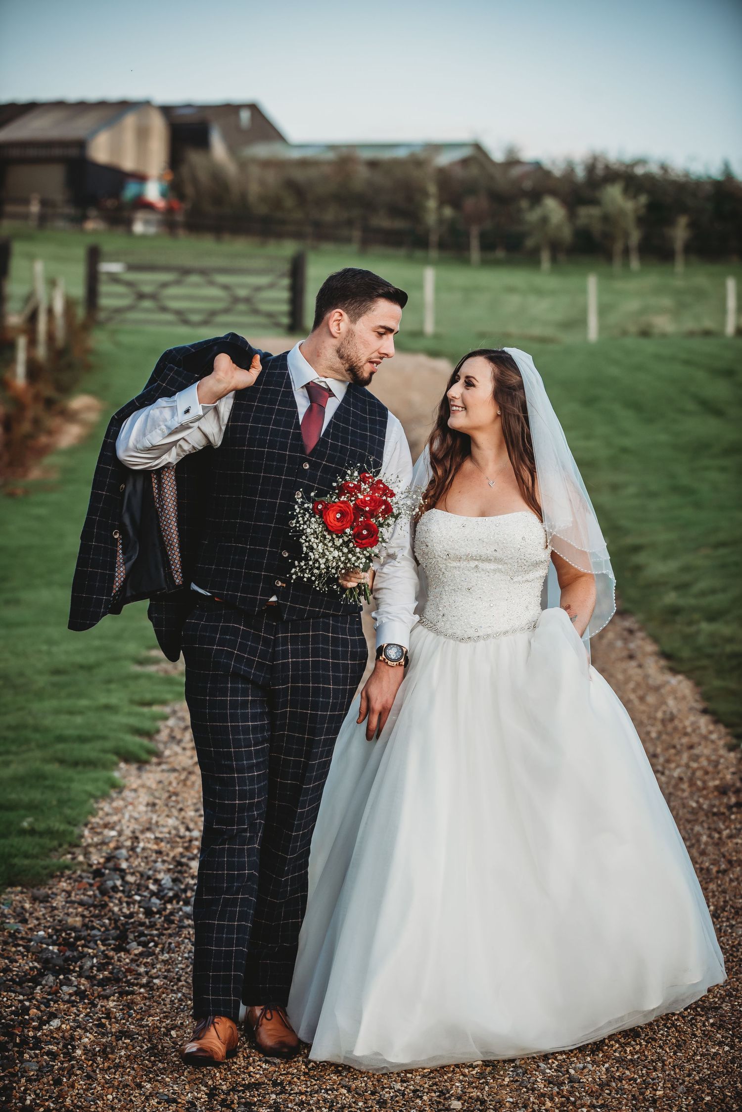 Newlyweds on rustic path in countryside setting with bride in white gown and groom in navy checkered suit.