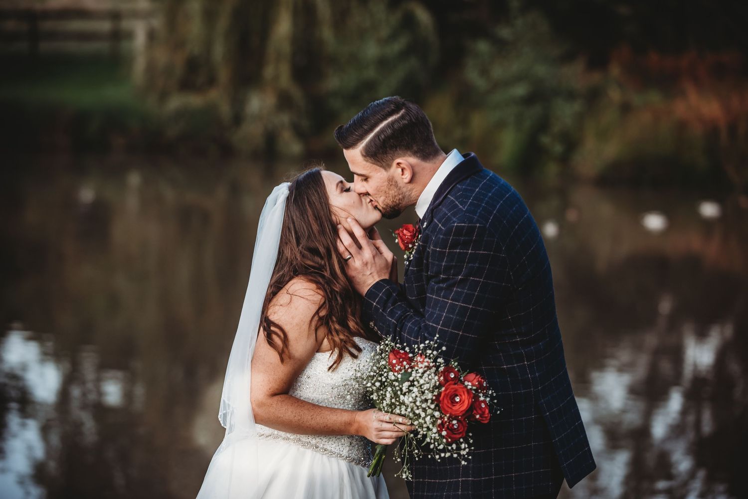 A romantic wedding couple shares a tender moment by a lake while holding a red rose bouquet.