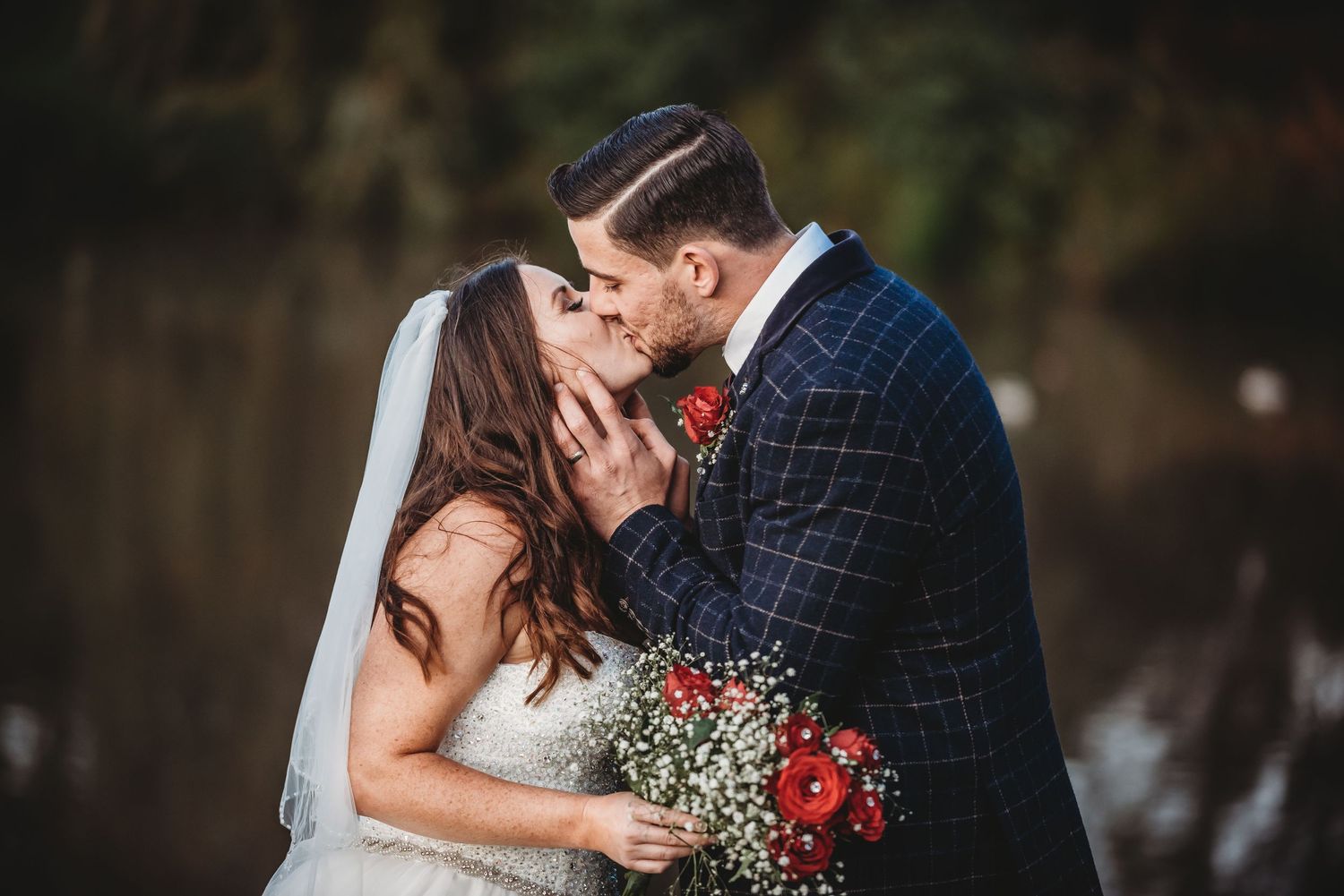 A romantic outdoor wedding scene with an elegant couple sharing a kiss while holding red floral bouquet.