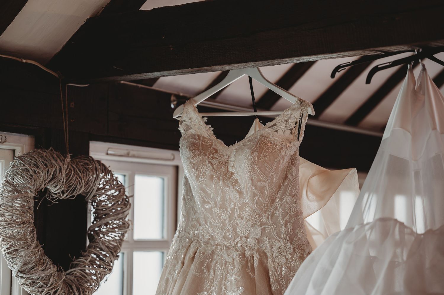 A white lace wedding dress hangs elegantly beside a decorative wreath in a rustic room with exposed beams.
