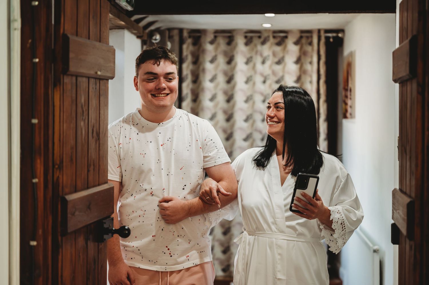 Two friends in white outfits smile while standing in a hotel hallway with cream-colored wallpaper and wooden accents.
