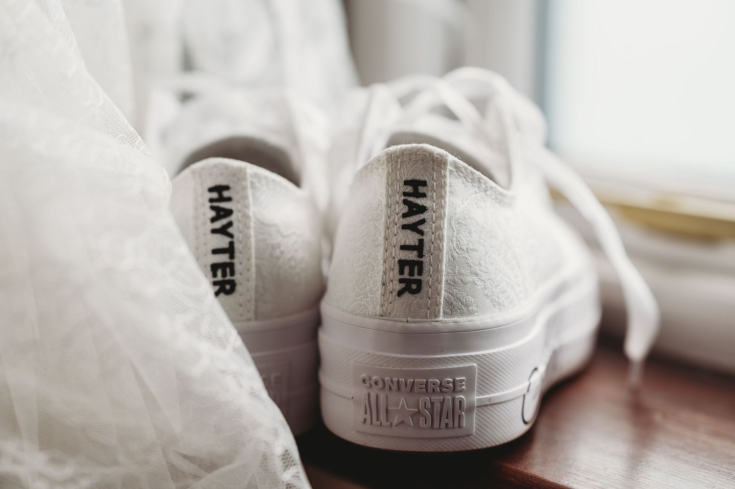 A pair of white Converse All Star sneakers sits on a wooden surface near delicate fabric.