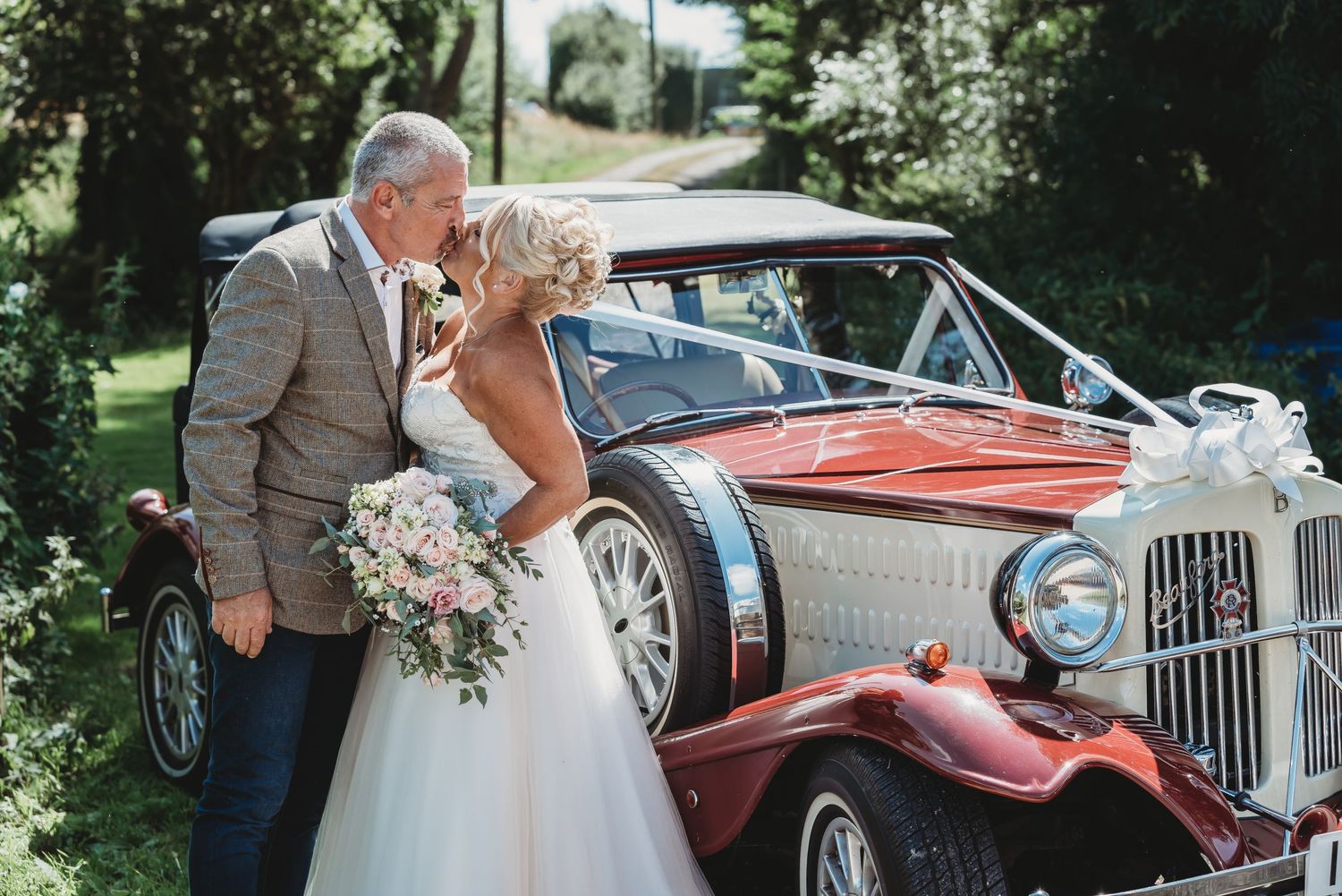 A classic vintage red car adorned with wedding decorations parked on a sunny day amidst greenery.