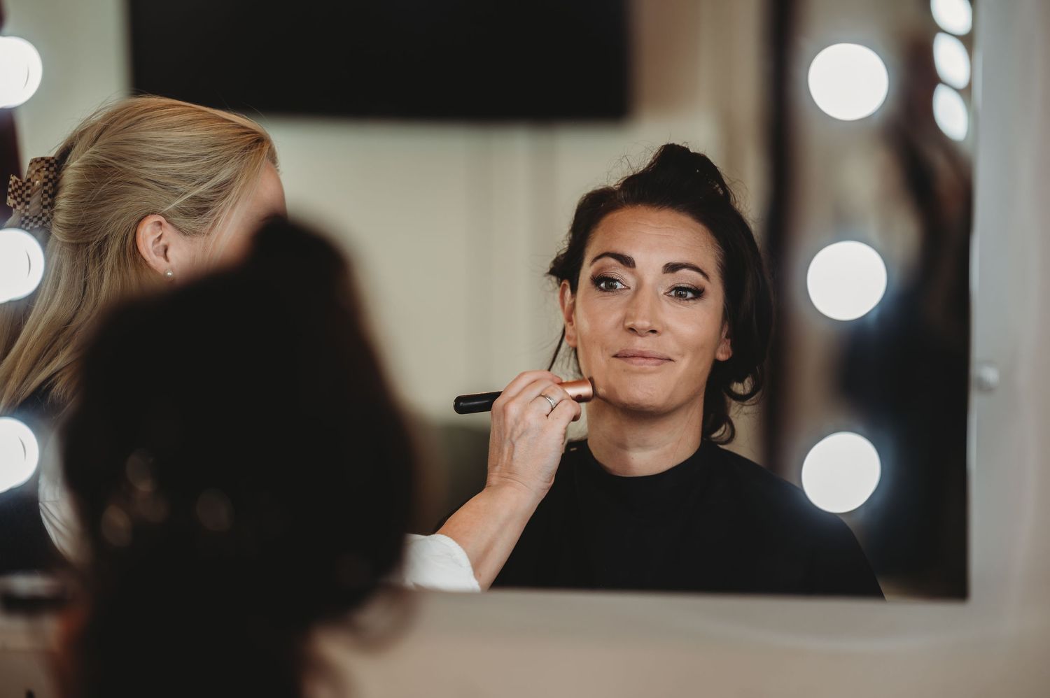 Professional makeup artist applies cosmetics on client in front of illuminated mirror during beauty session.
