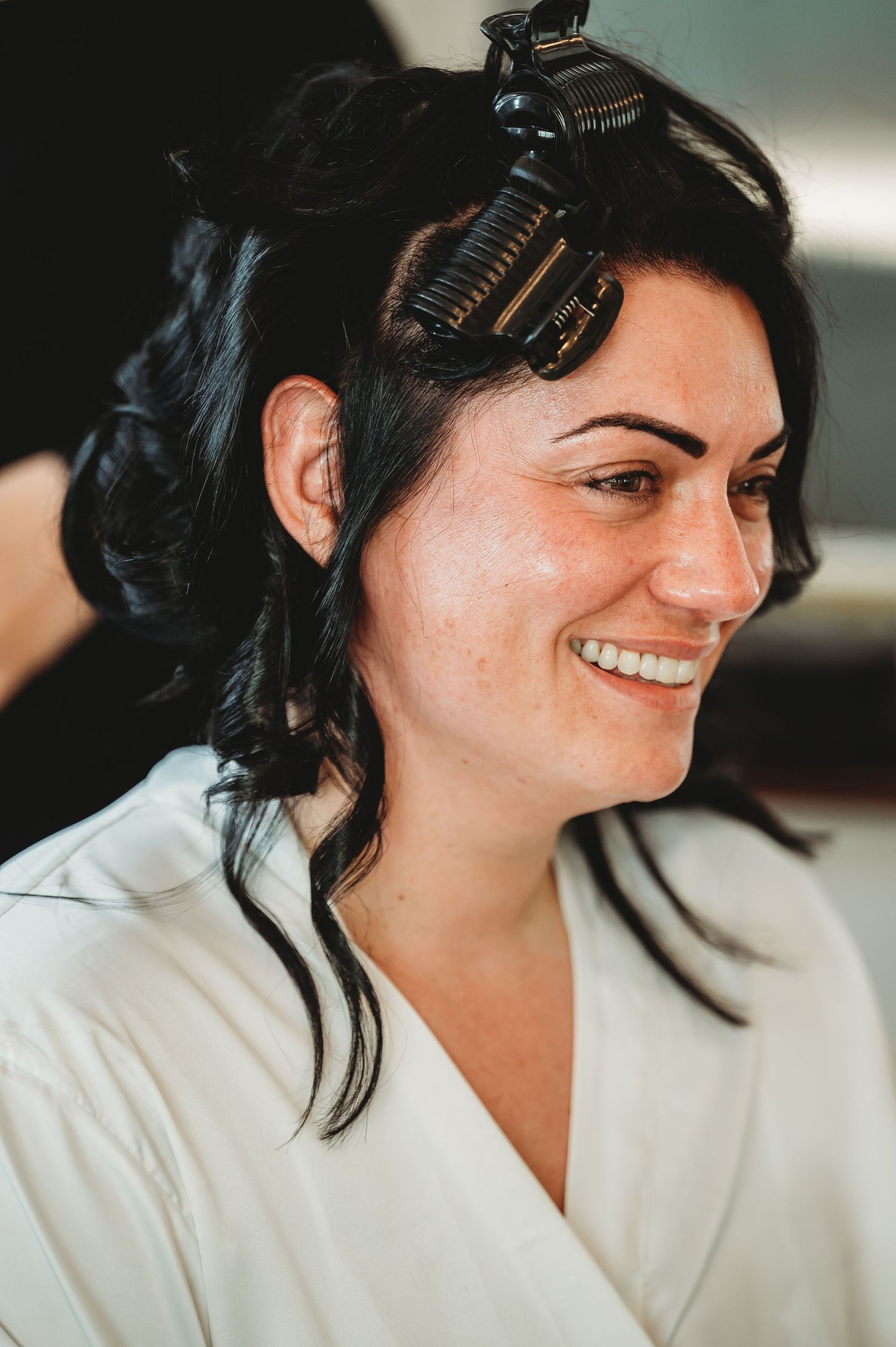 A candid shot of someone getting ready with hair in curlers, wearing a white robe and smiling warmly.