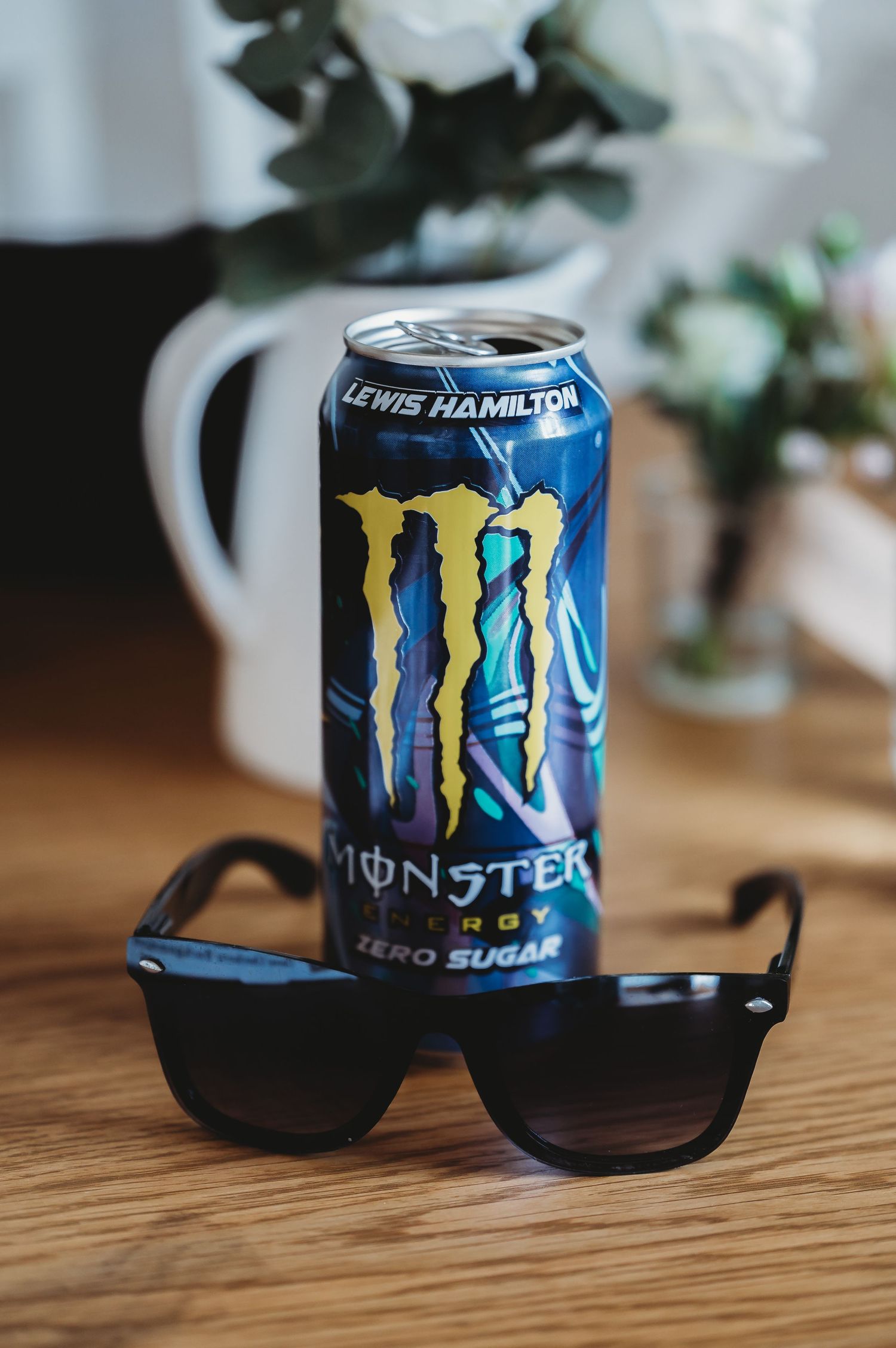 A blue Monster Energy drink can sits next to black sunglasses on a wooden surface with a white mug in the background.