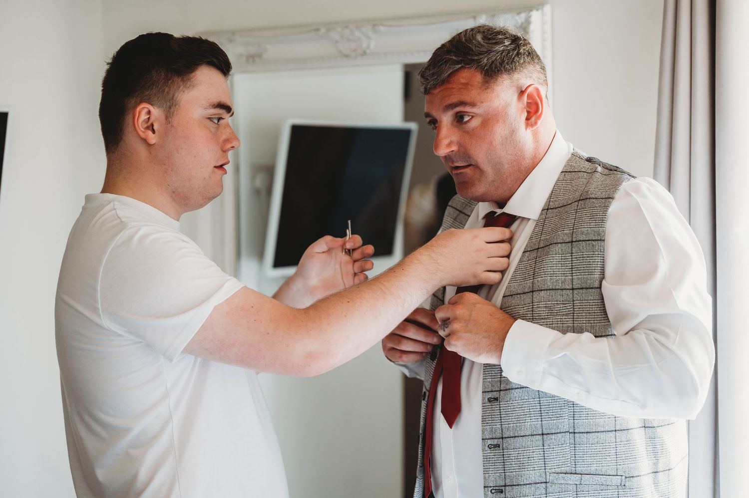 Two people in white shirts getting ready, with one helping adjust a red tie on the other's grey checkered vest.