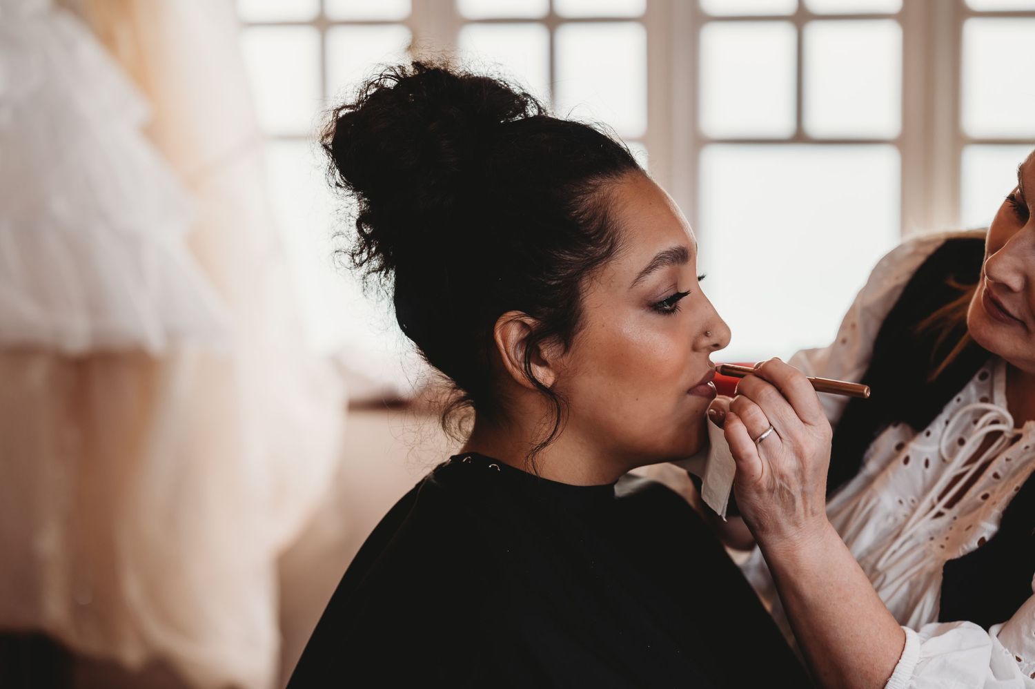 Makeup artist applies lipstick on client during professional beauty session in natural window light.