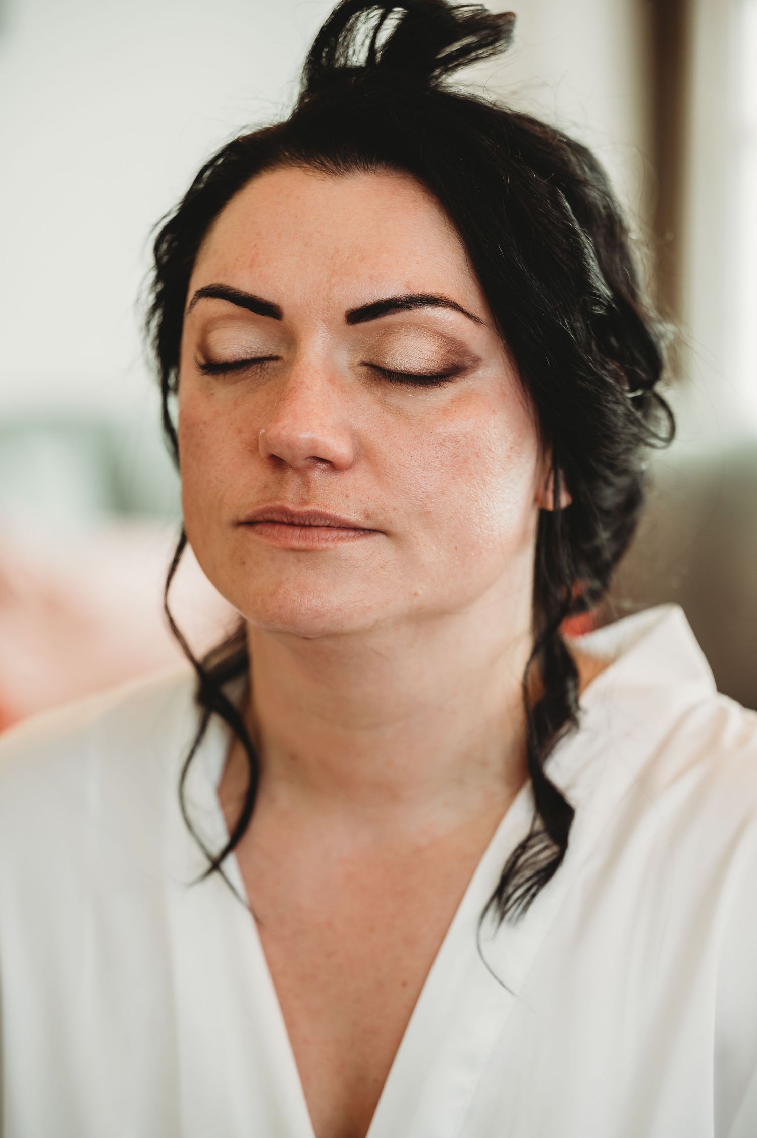 A close-up portrait of someone with closed eyes, dark hair in a messy bun, wearing a white robe.