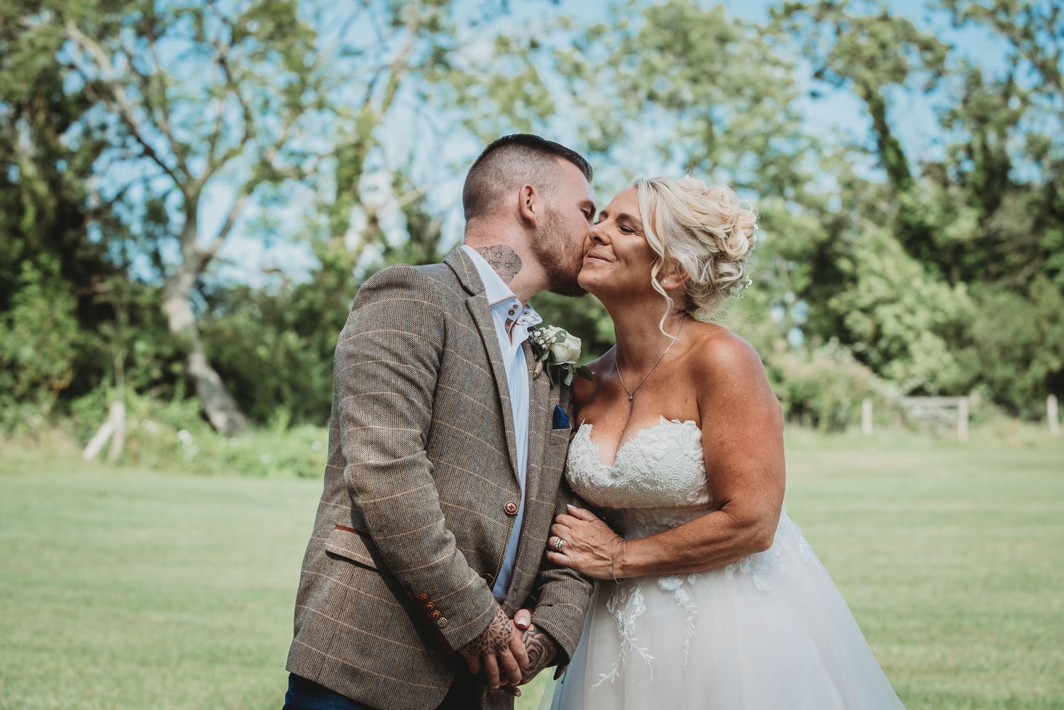 A couple embraces and kisses on their wedding day outdoors on a green lawn with trees in the background.