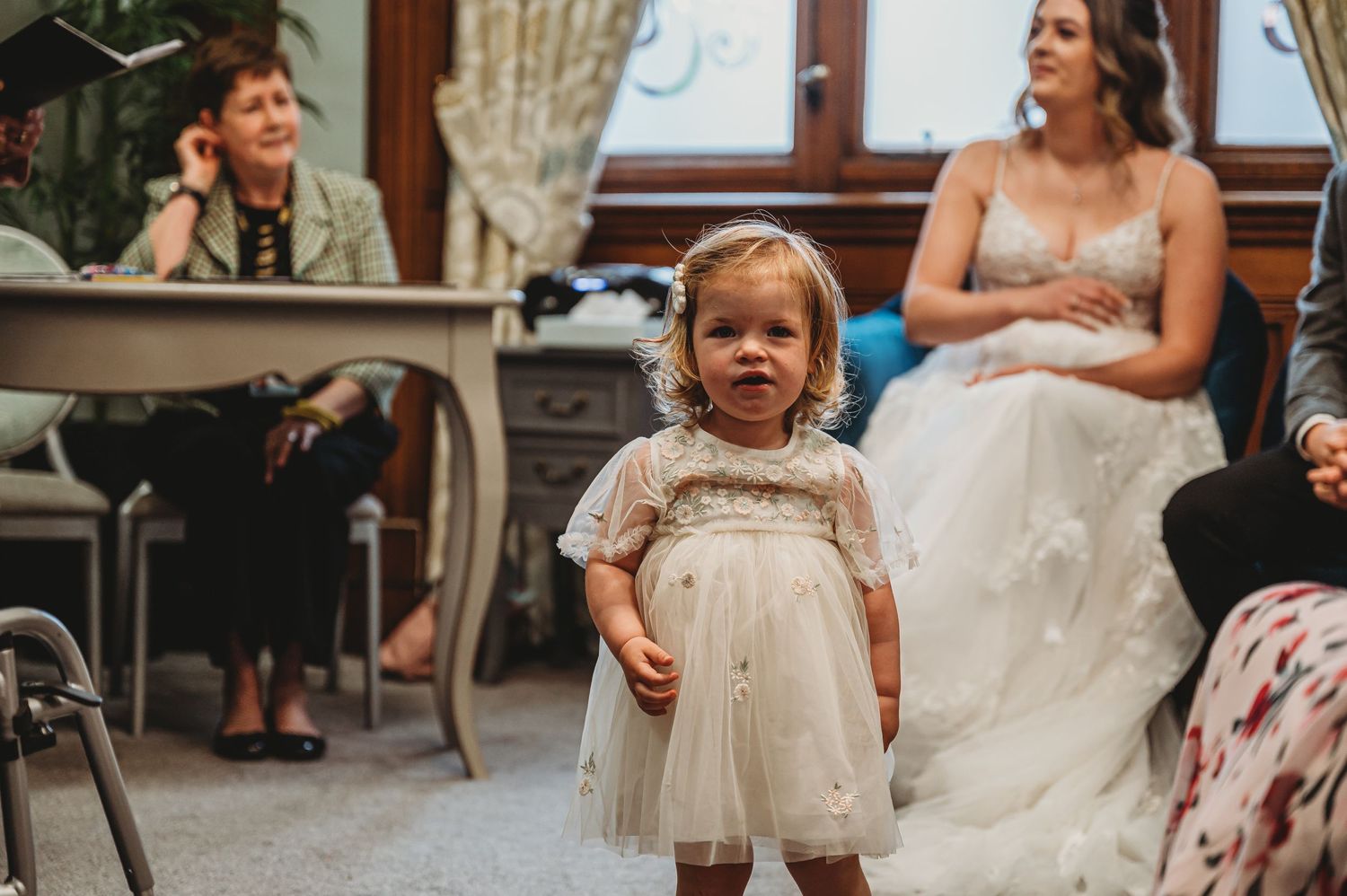 Small child in white dress stands on dance floor during wedding reception with guests seated in background.
