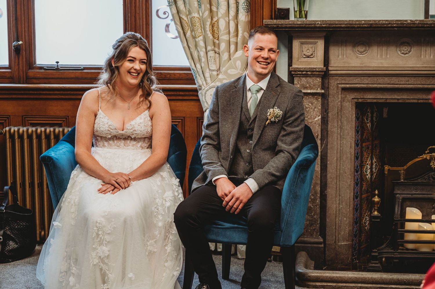 A newlywed couple sits together on blue velvet chairs in an elegant room with a fireplace and wooden details.