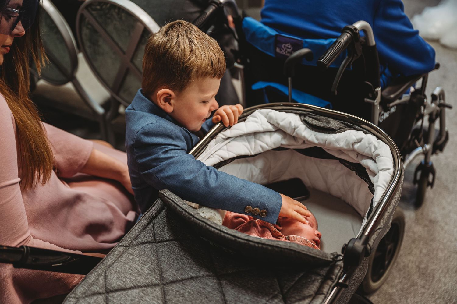 Young child in blue sweater reaches into gray stroller to grab something at a store or shopping area.