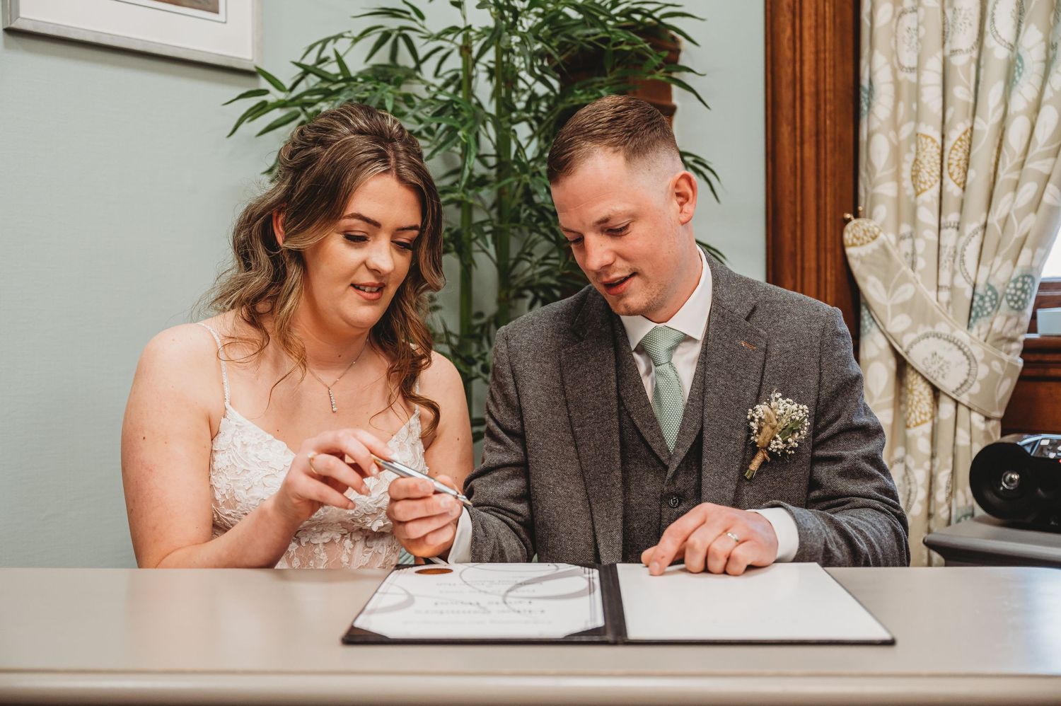 Two people at a table signing wedding documents while the bride examines her ring during a marriage ceremony.
