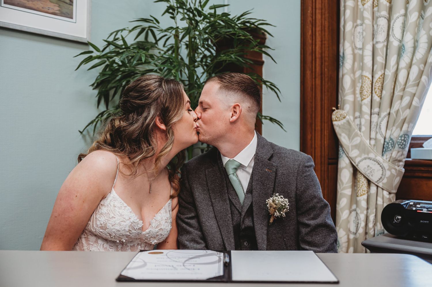 A romantic newlywed couple shares a kiss at their wedding ceremony while sitting at a table with documents.