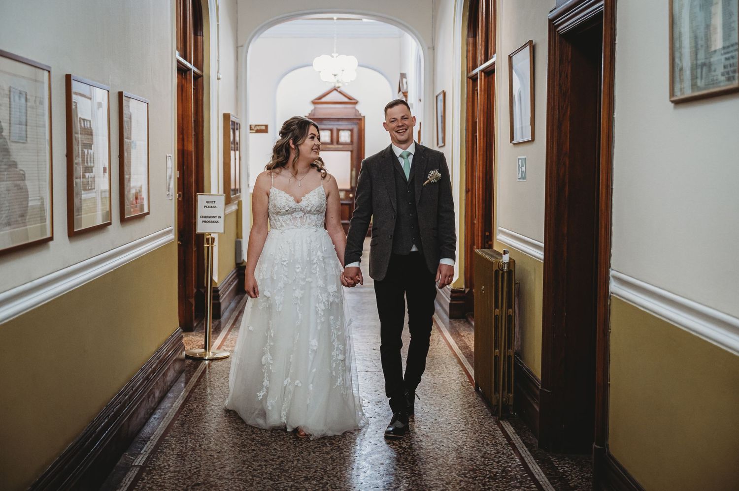 Couple walks hand in hand down an elegant hallway with arched ceiling and framed artwork on yellow walls.