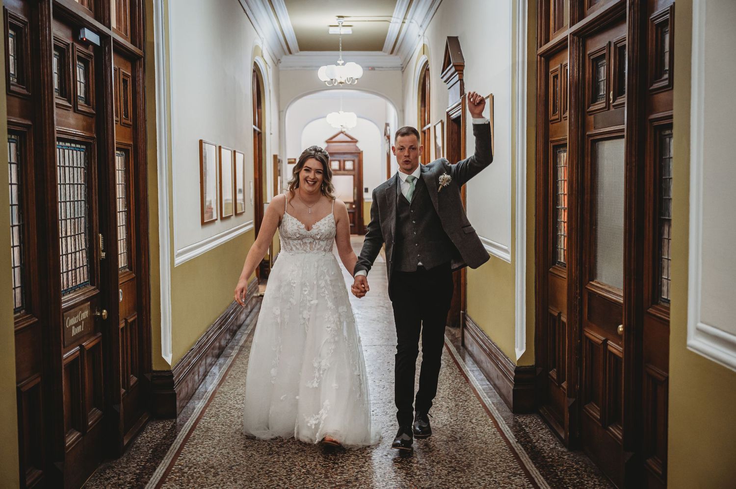 A joyful couple celebrates in an elegant hallway with ornate wooden doors and yellow walls on their wedding day.